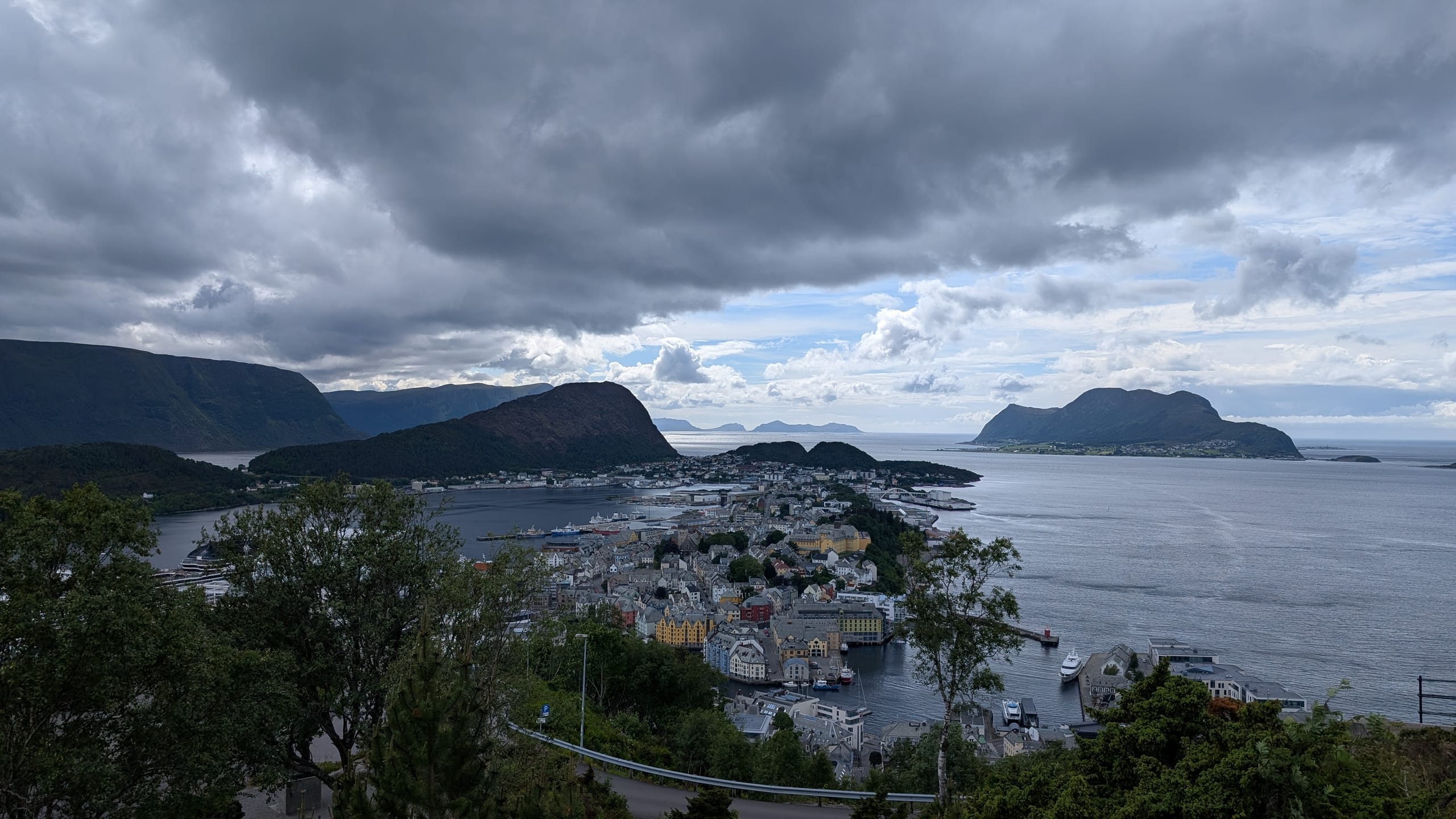 Ålesund spread across fjord islands under heavy storm clouds, seen from the green hillside above town