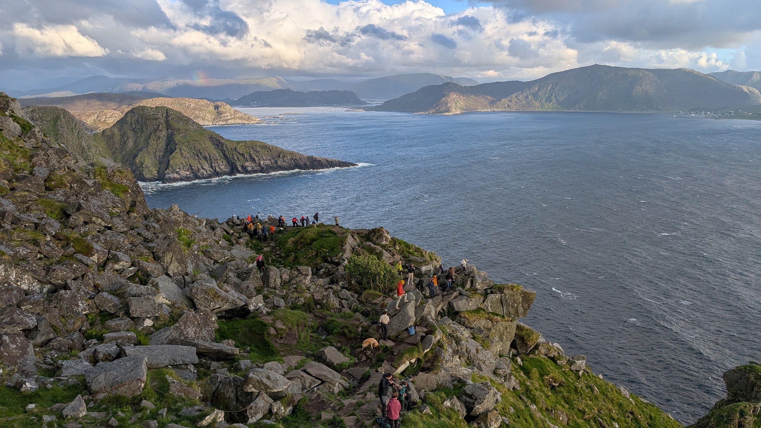 Hikers on a rocky green clifftop on Runde looking out over windy blue sea toward islands, with a faint rainbow under broken cloud