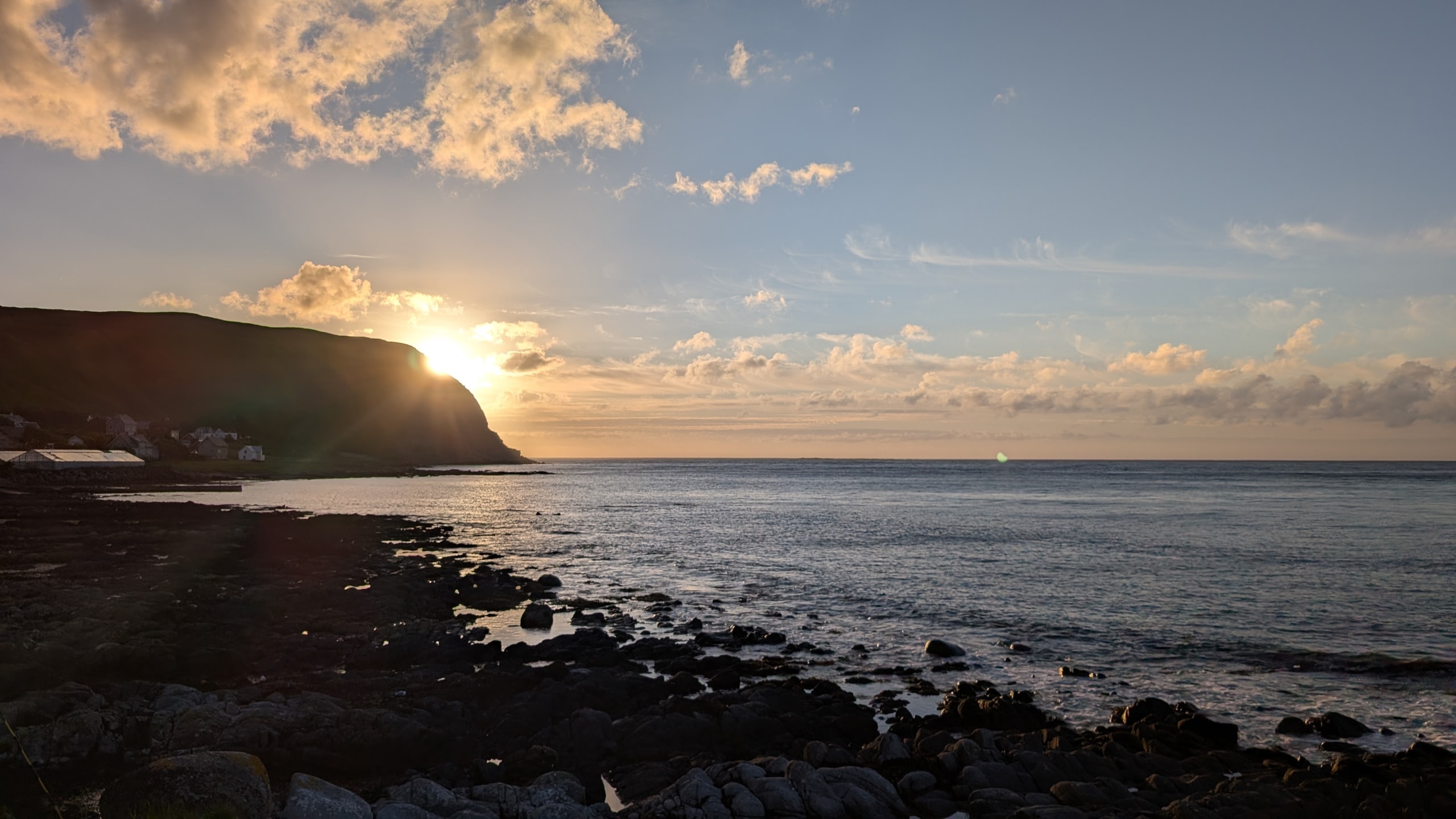 Sun setting behind a dark coastal headland near Ålesund, with golden light on calm water and a rocky shoreline in the foreground