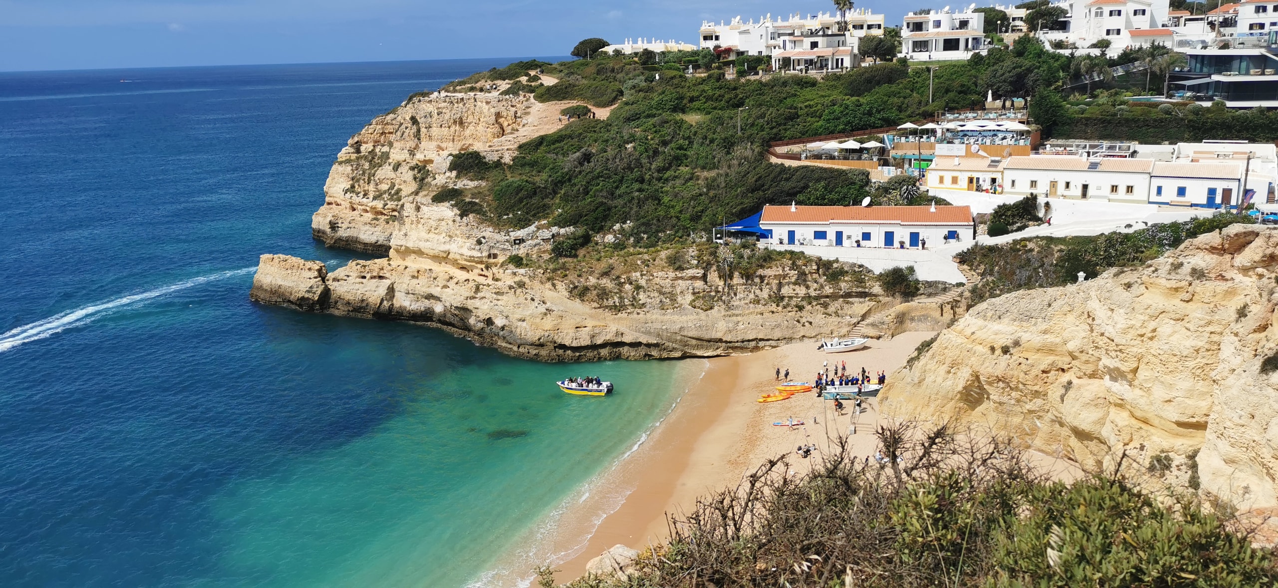 A small sandy cove under ochre cliffs with white houses, kayaks on the beach, and a boat in turquoise water near Carvoeiro