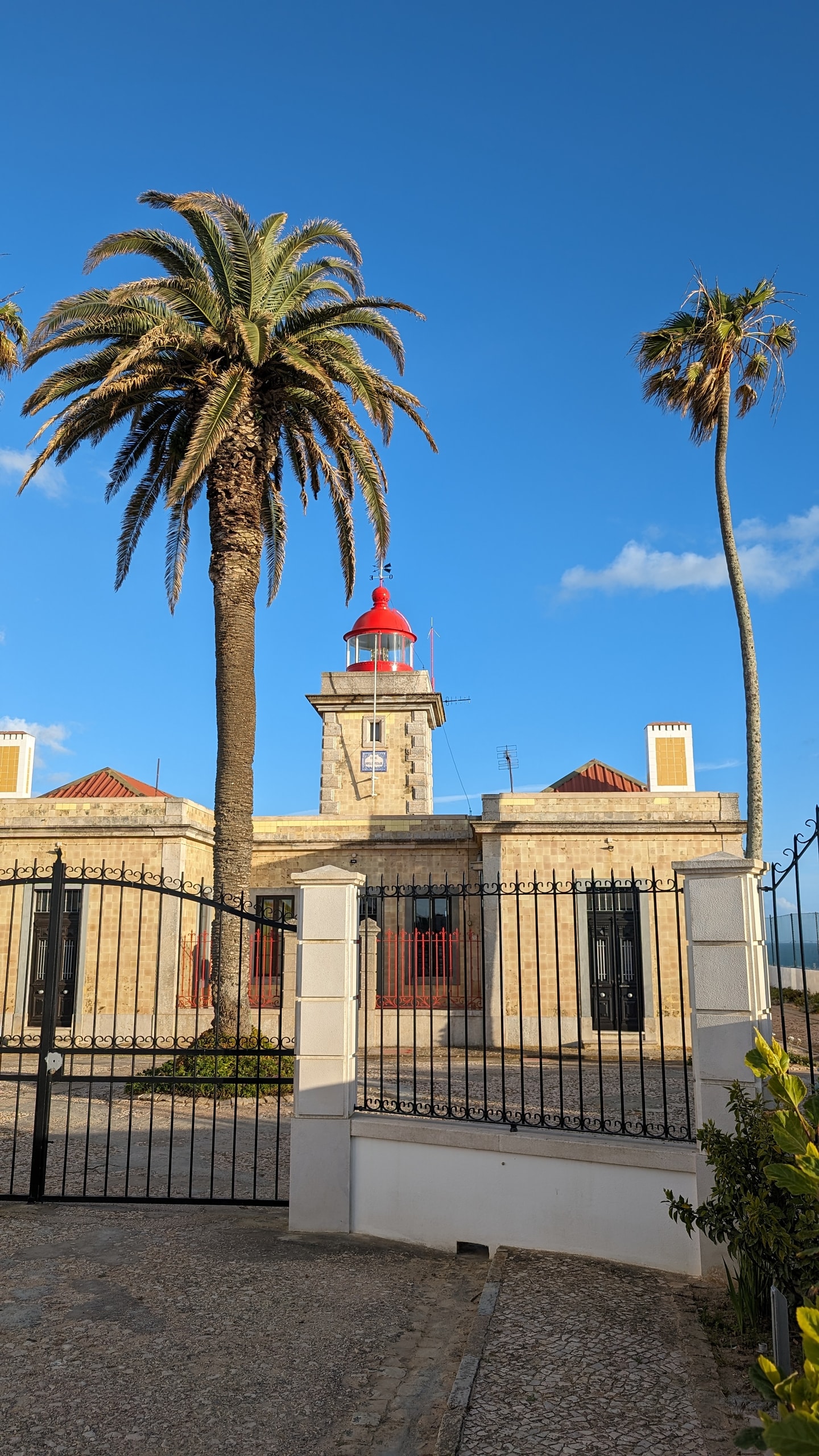 The square stone Ponta da Piedade lighthouse with a red lantern, palm trees, and a black iron gate on cobblestones
