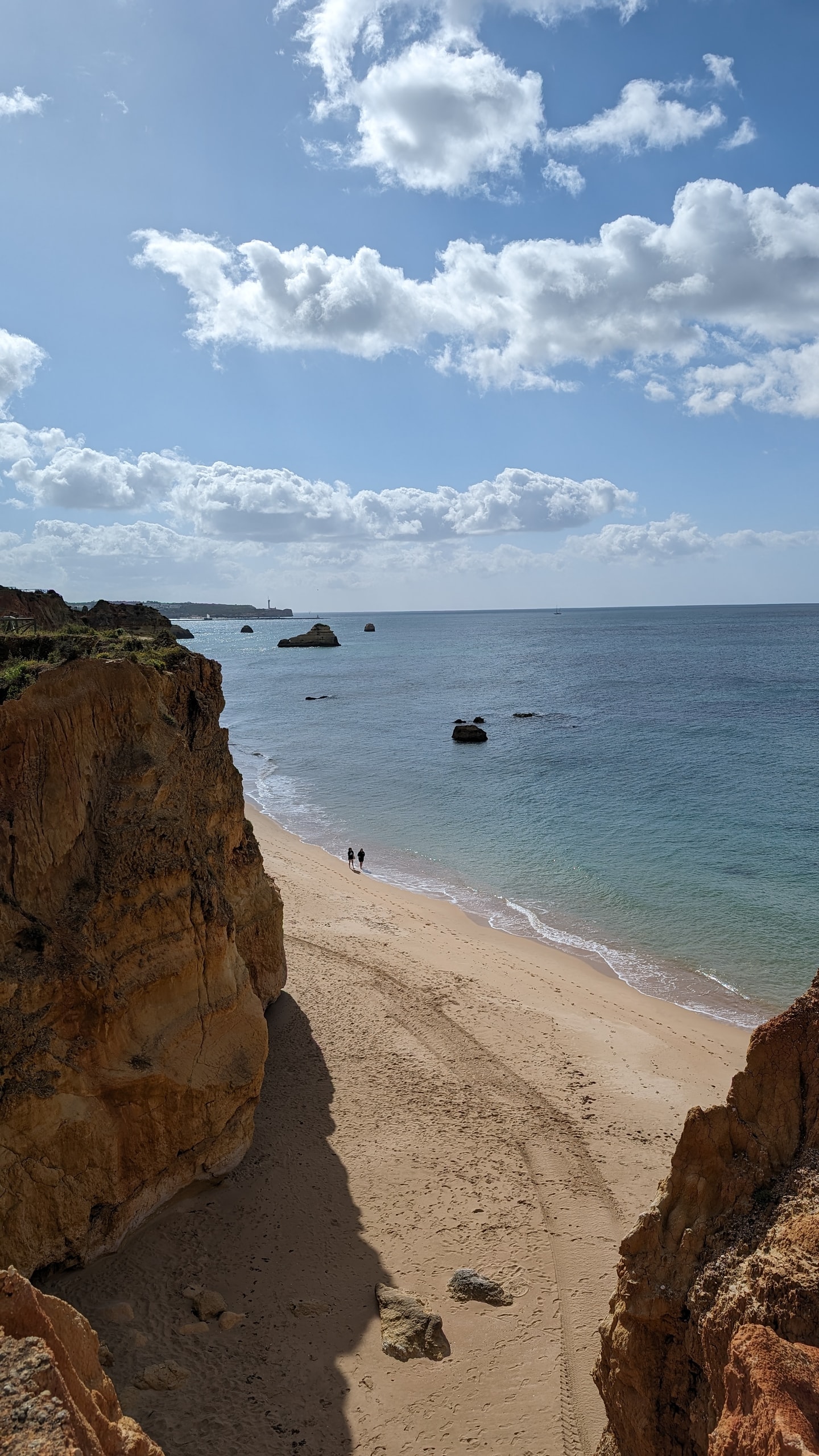 Looking down from ochre cliffs onto Praia da Rocha’s wide sand, turquoise shallows, and two small figures walking the shoreline