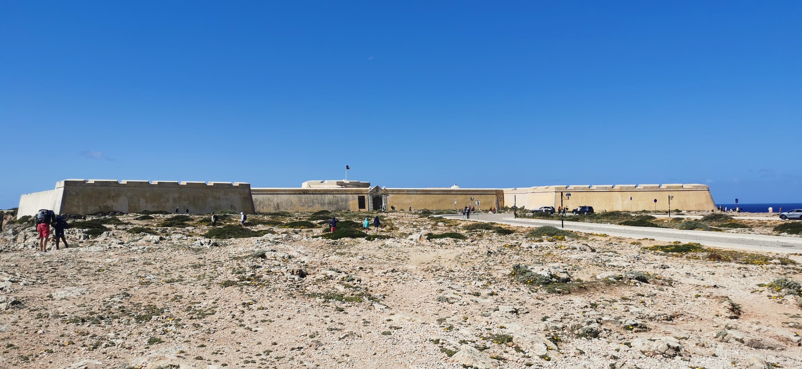 The long pale stone walls of Sagres Fortress across a rocky plateau under a clear blue sky, with the Atlantic visible to the right