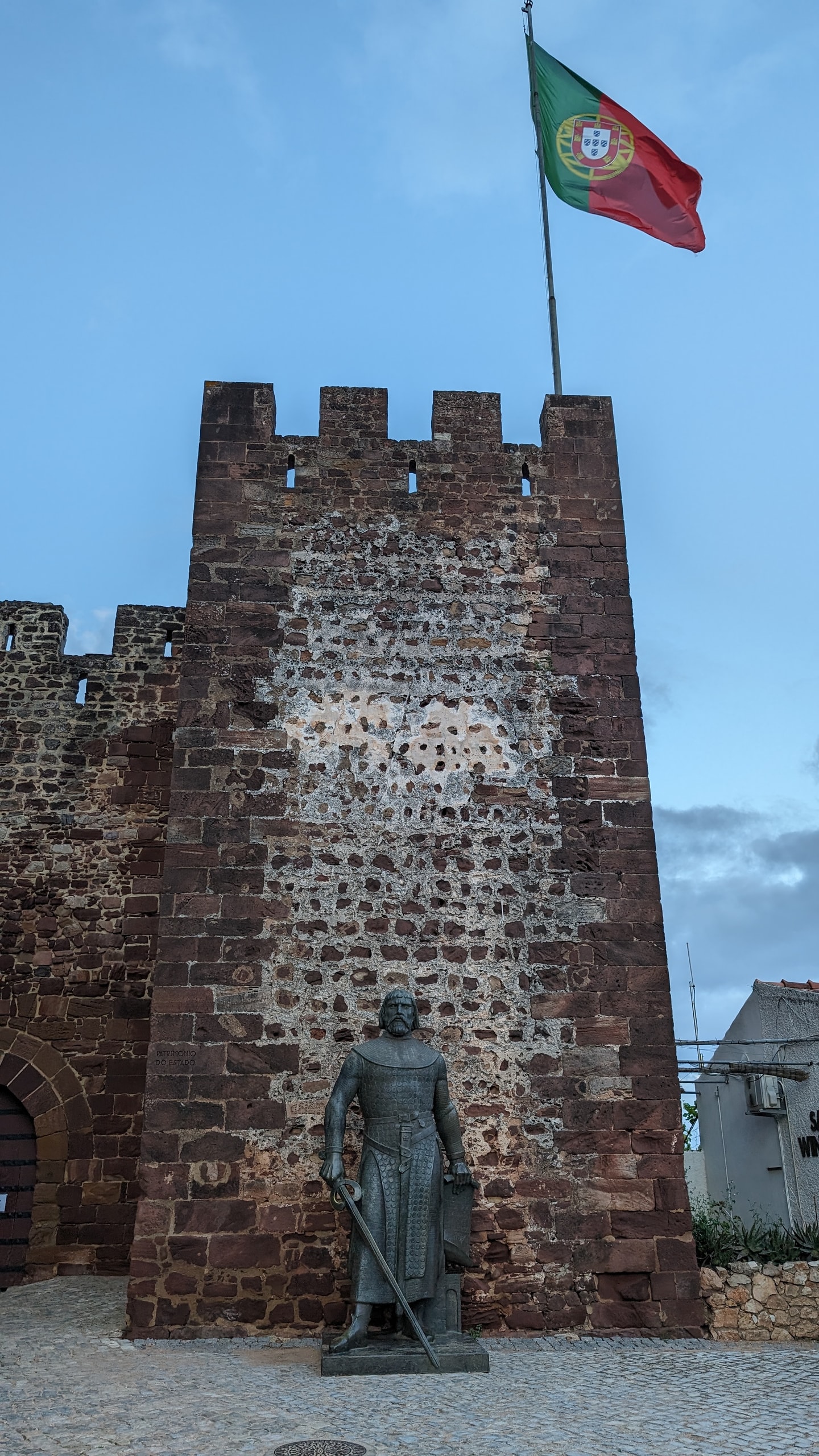 Bronze statue of King Sancho I in chainmail before the red sandstone tower of Silves Castle, with the Portuguese flag on the battlements