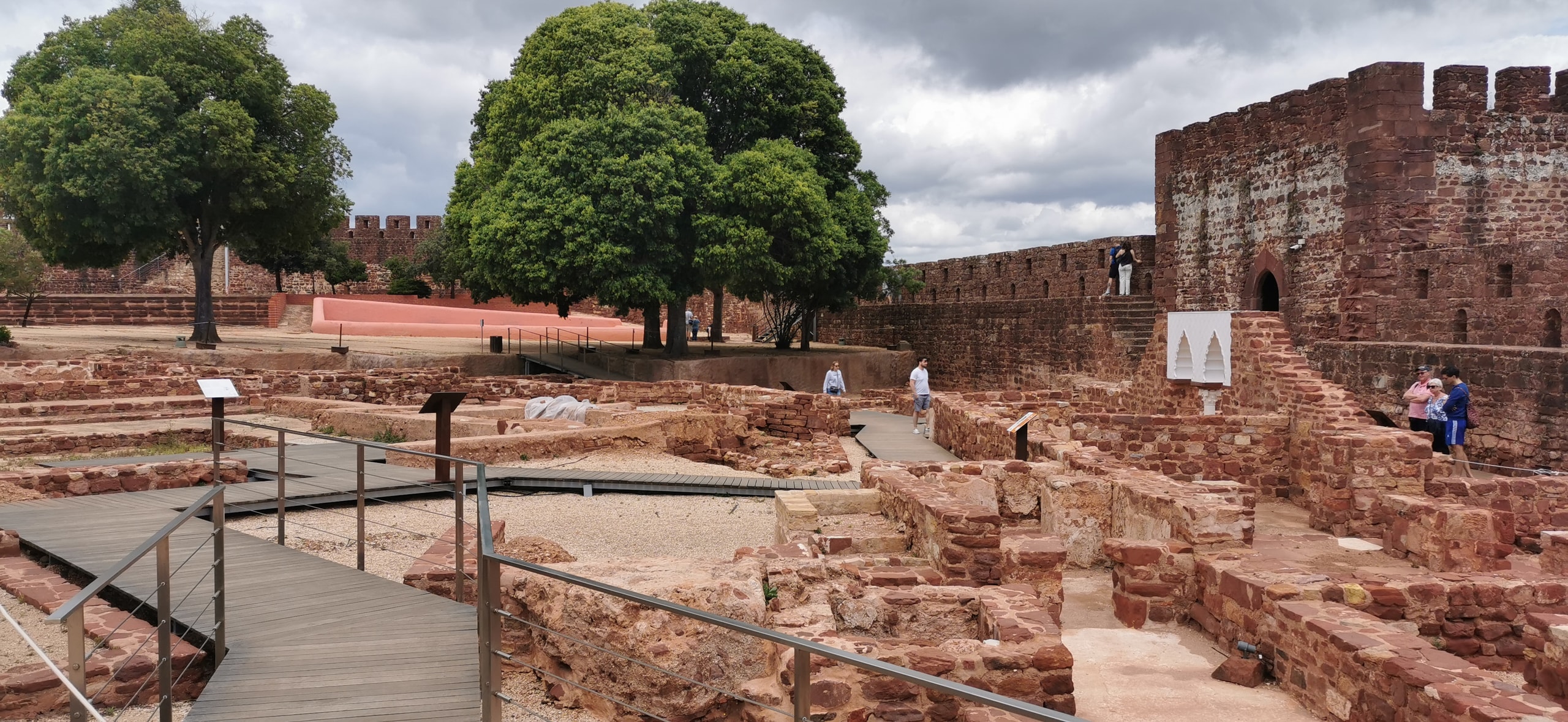 Wooden visitor walkways through excavated red sandstone foundations inside Silves Castle, with a square tower and large trees