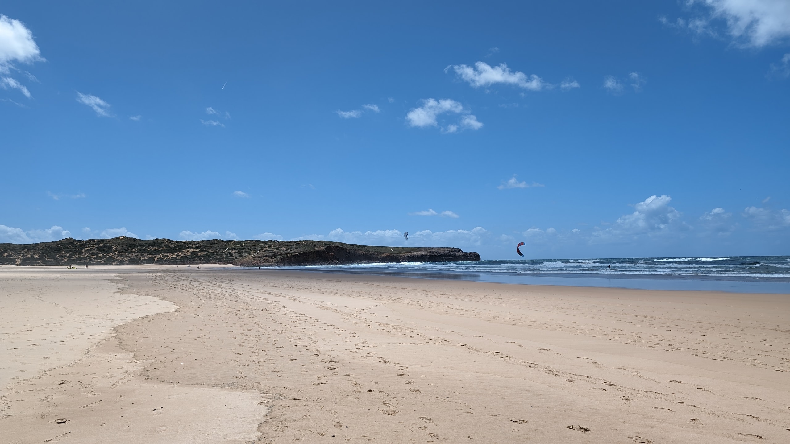 A wide Vicentine Coast beach of pale sand and white surf under a clear sky, with kitesurfers in the distance
