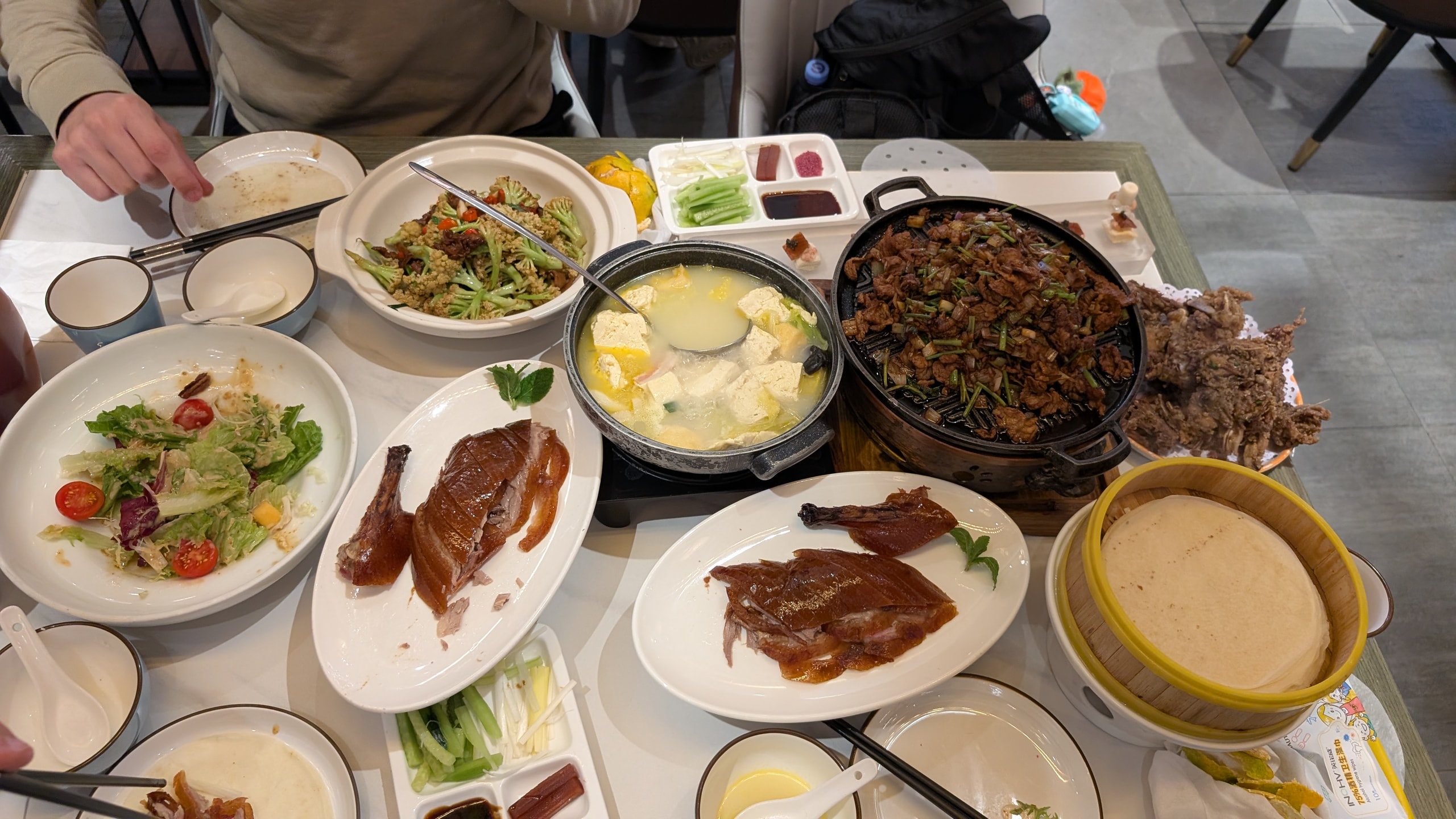 A Beijing dinner table covered with sliced roast duck, pancakes, vegetables, tofu soup, and shared meat dishes