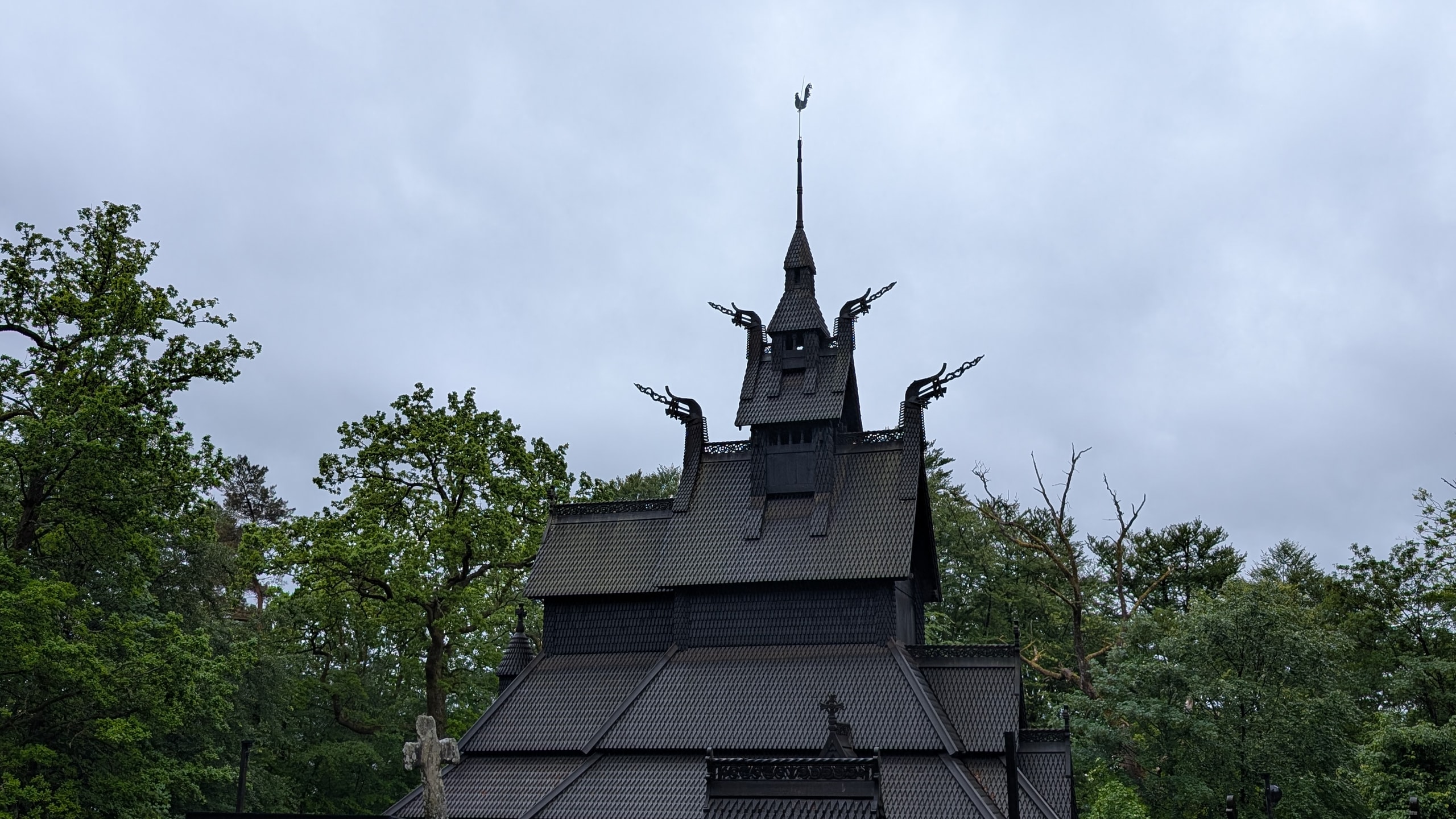 Dark tarred Fantoft Stave Church with dragon-head gables and a tall spire, set among green trees under a grey sky
