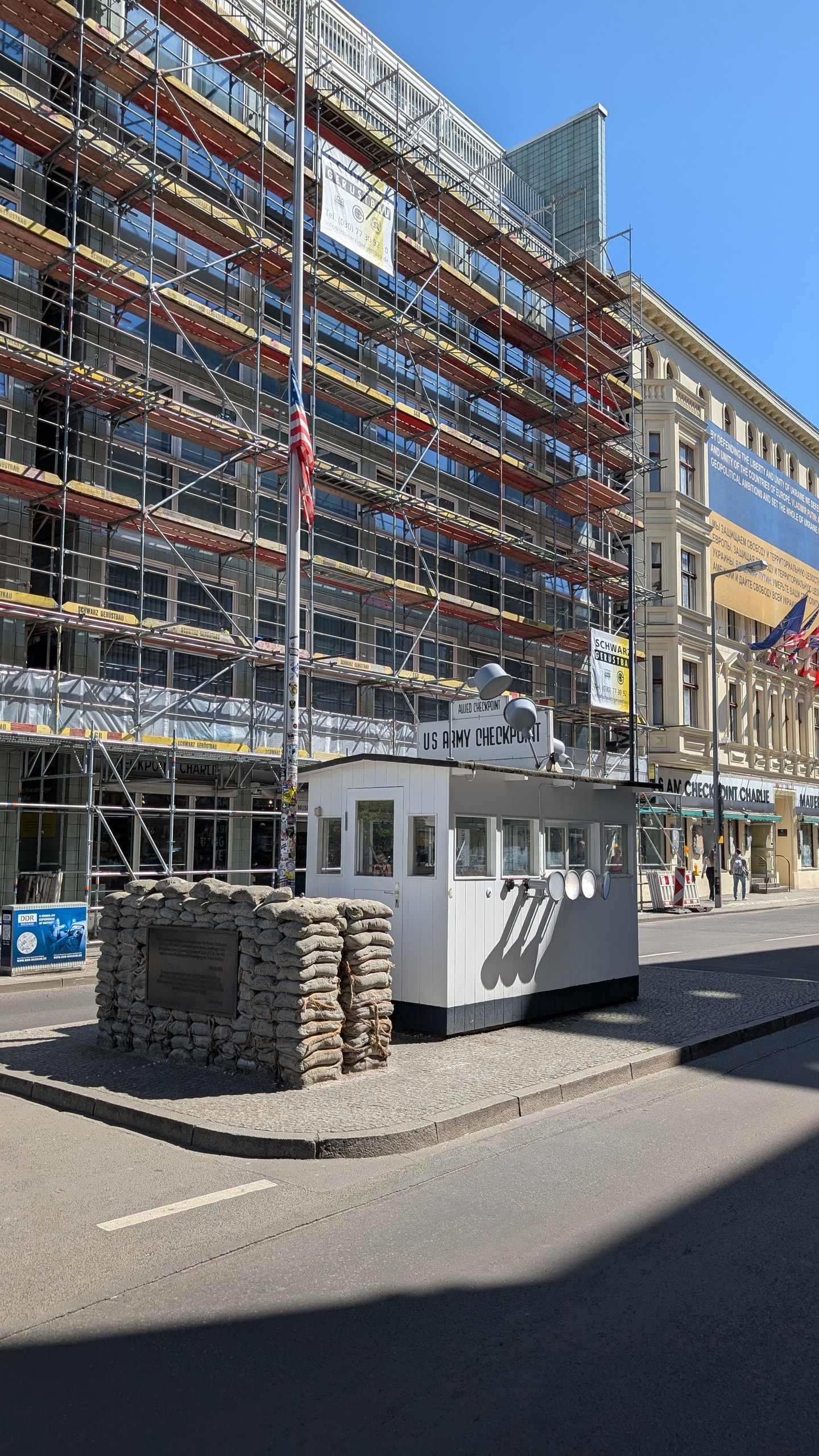 The replica US Army checkpoint hut at Checkpoint Charlie with stacked sandbags and a flag, in front of a building covered in construction scaffolding