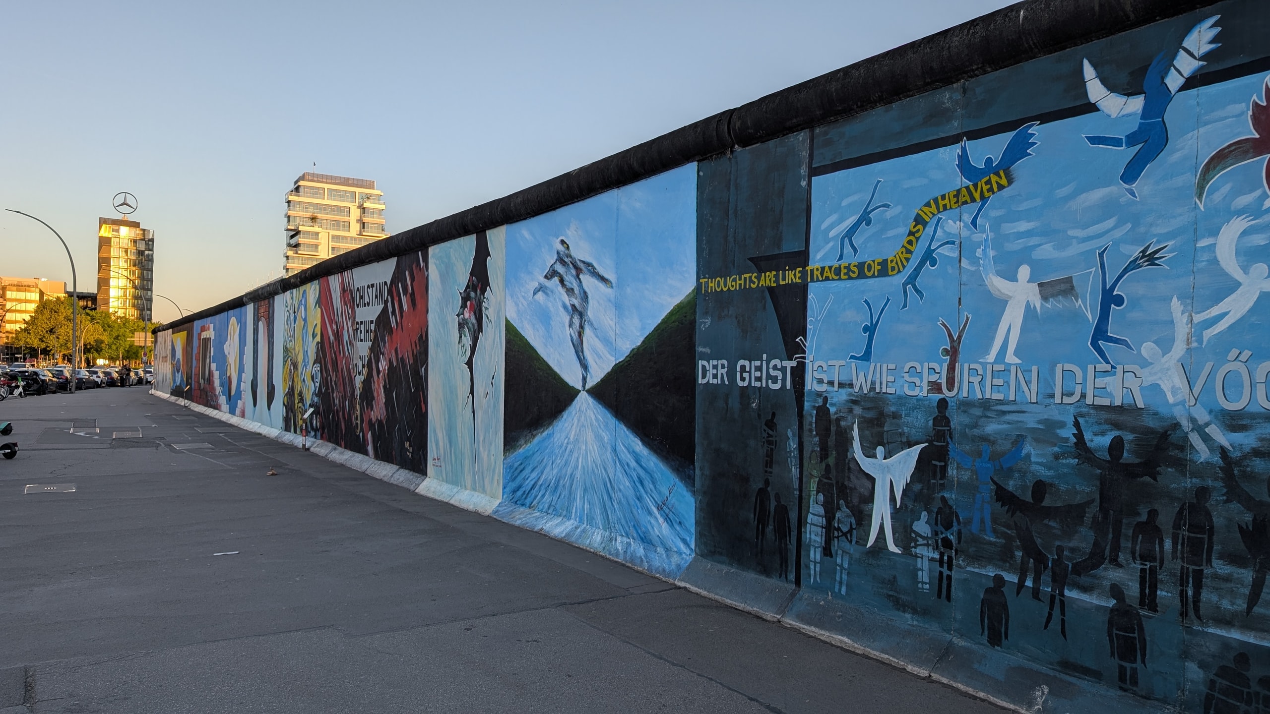 A long painted stretch of the East Side Gallery on Mühlenstrasse in low evening sun, with murals of birds and running figures