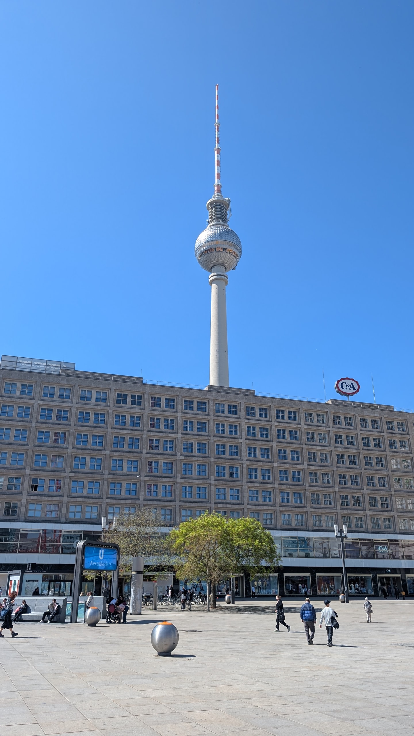 The Fernsehturm television tower rising behind the long facade of Alexanderplatz under a clear blue sky, with people crossing the empty square
