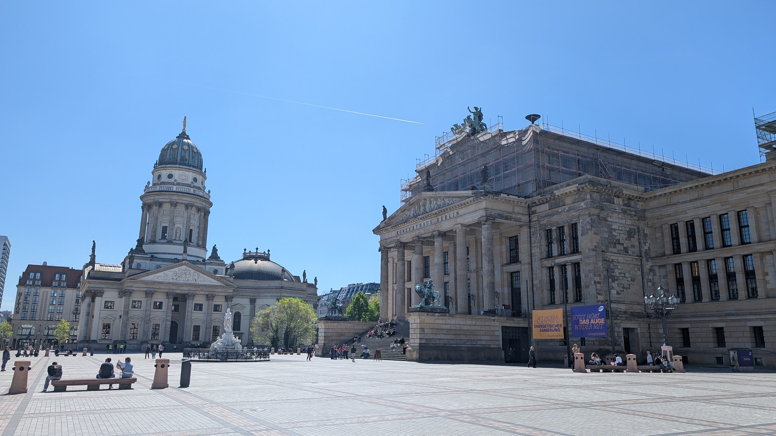 Gendarmenmarkt square in Berlin with a domed cathedral on the left and the columned Konzerthaus under scaffolding on the right, benches on the empty paving