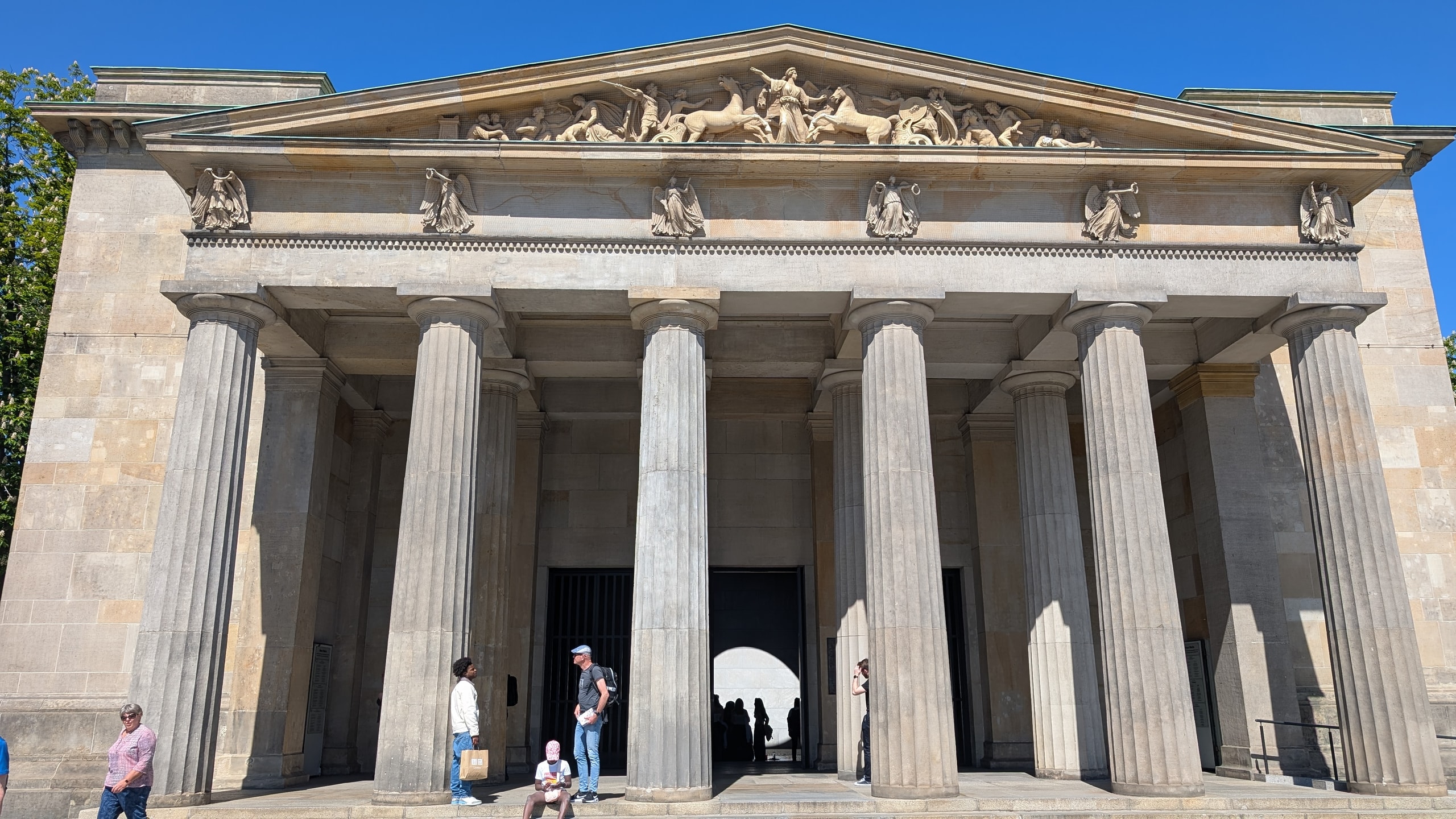 The Neue Wache in Berlin, a neoclassical guardhouse with heavy Doric columns and a sculpted pediment, visitors standing on the steps in front of the dark open doorway