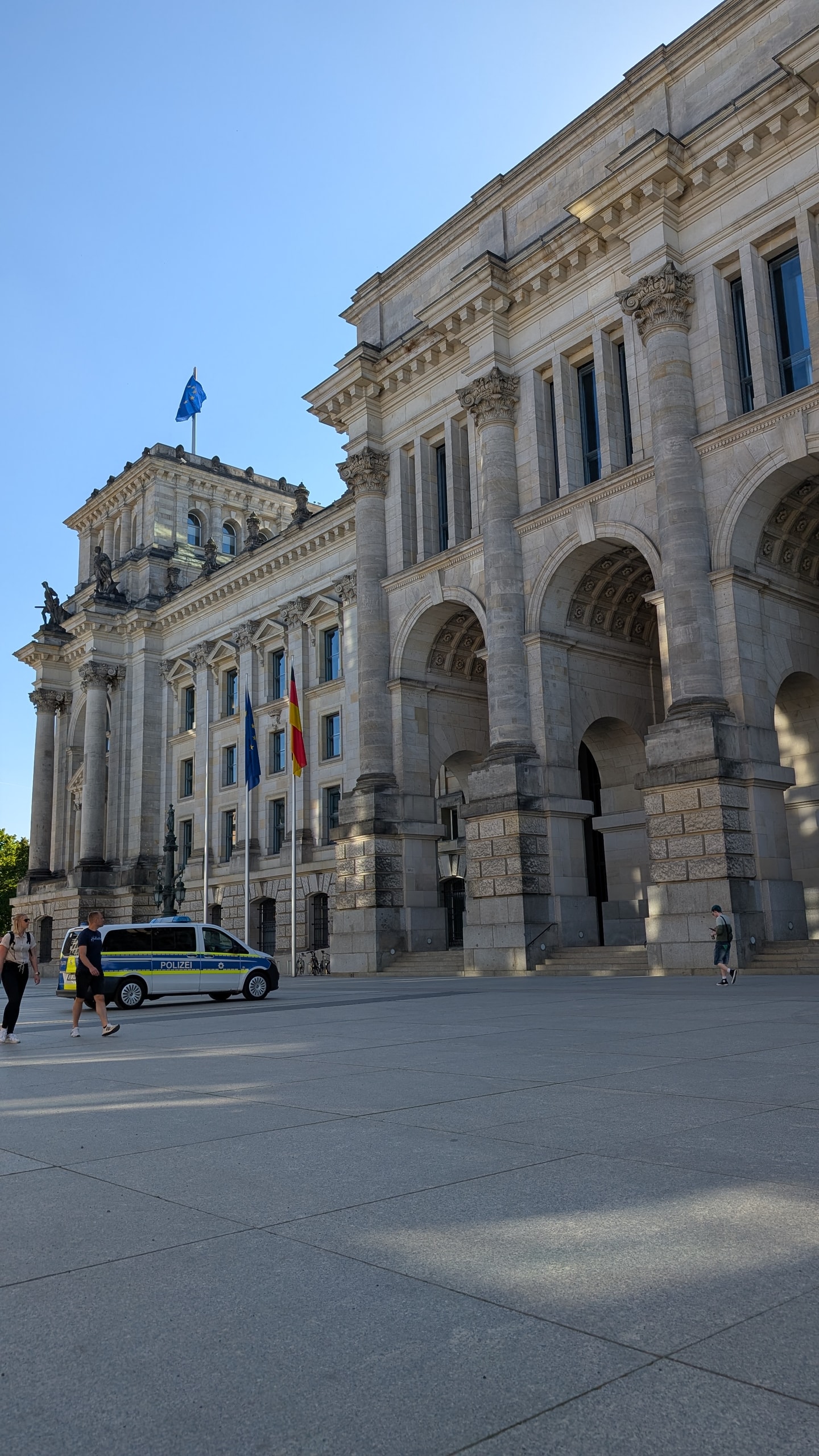 The stone arches and columns along the side of the Reichstag building in Berlin under afternoon sun, with the German and EU flags flying and a police van parked outside