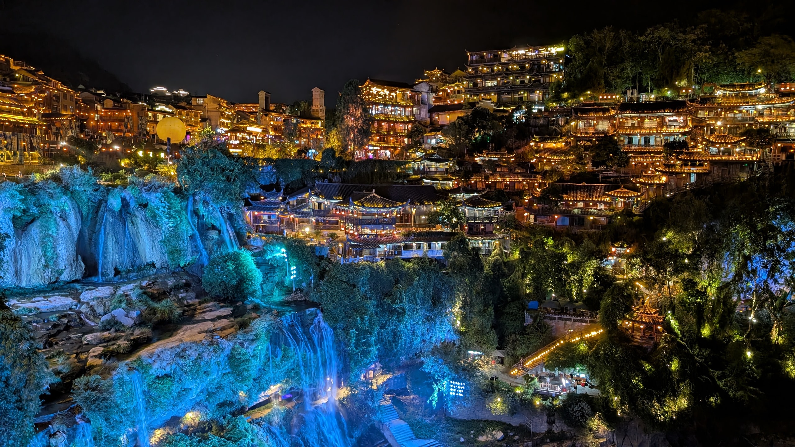 Furong Ancient Town at night with lit wooden buildings cascading down a cliff above a blue-lit waterfall