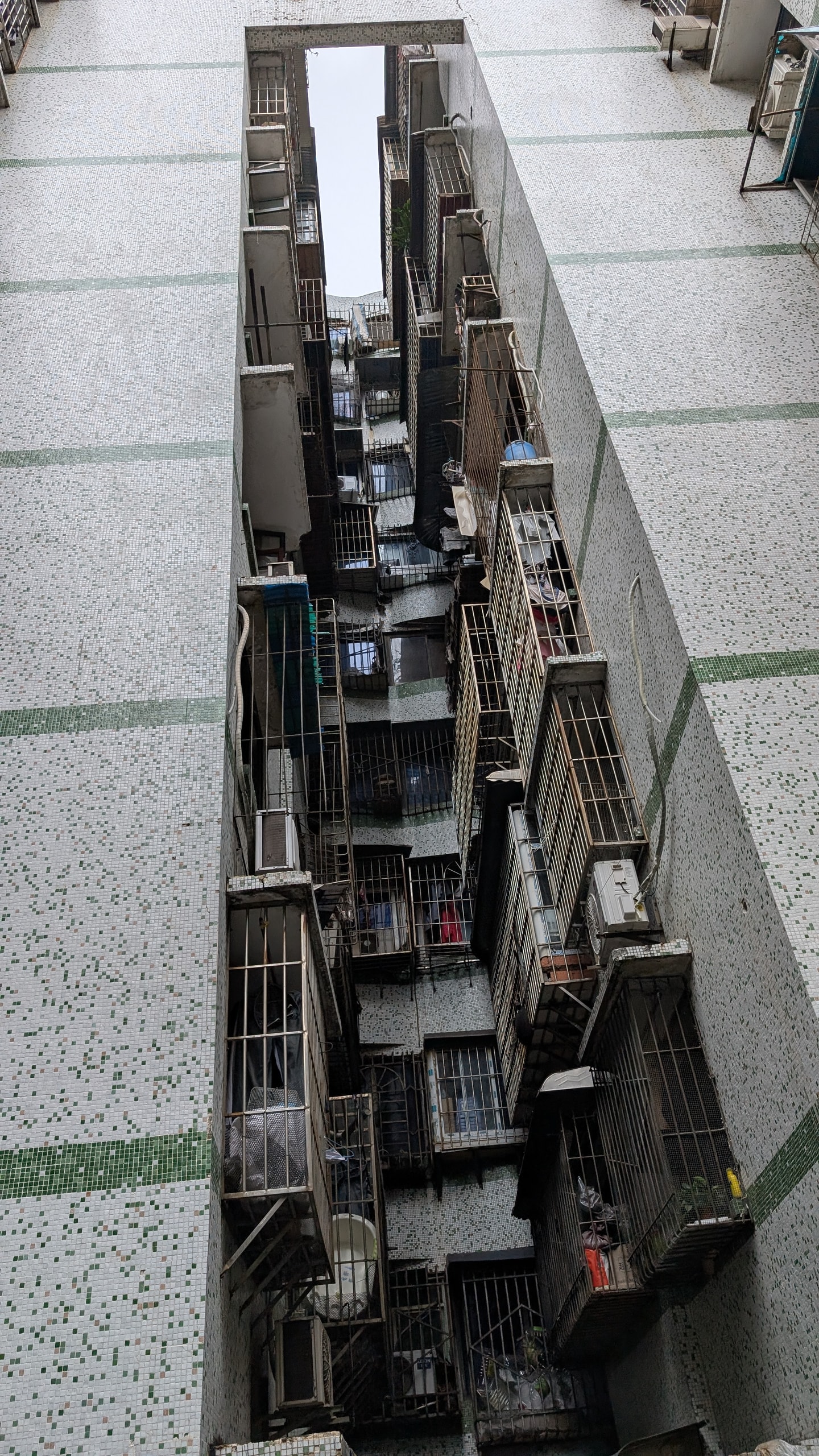 Looking straight up a narrow airwell between two tall tiled apartment buildings with window cages and laundry