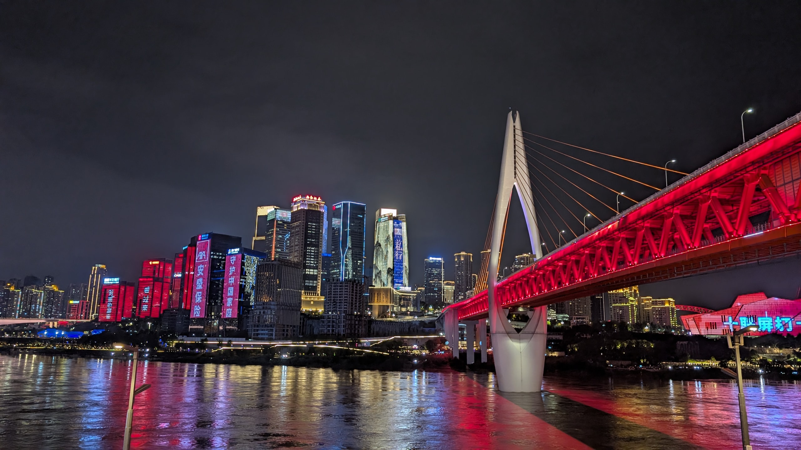 Chongqing night skyline with a red-lit cable-stayed bridge and skyscraper LED displays reflecting in the river
