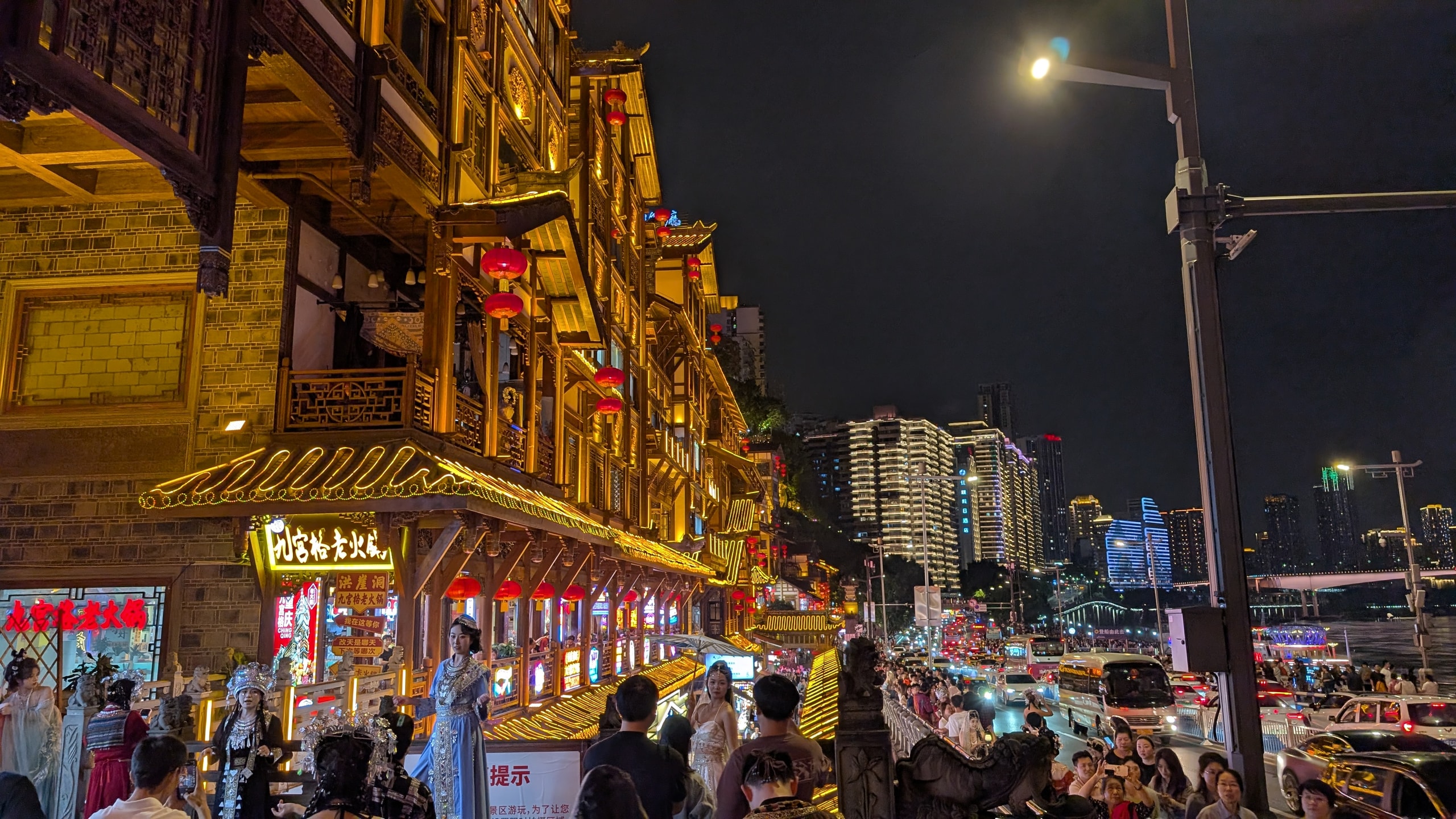 Crowds on the walkway outside Hongya Cave at night, with lit traditional facades, red lanterns, and traffic on the road below