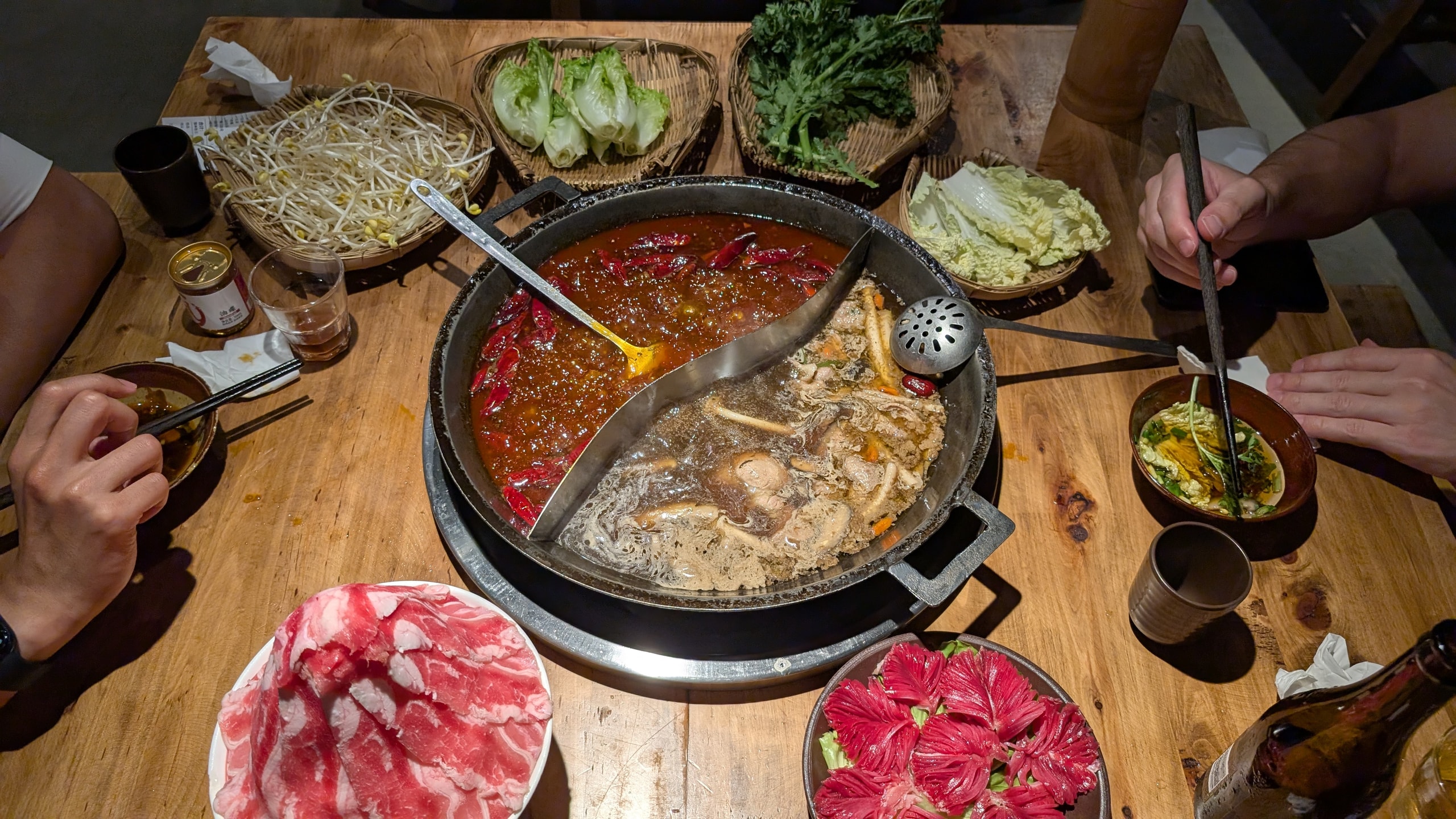 Overhead view of a split Chongqing hotpot with red chili broth and mild broth, surrounded by beef, greens, and dipping bowls