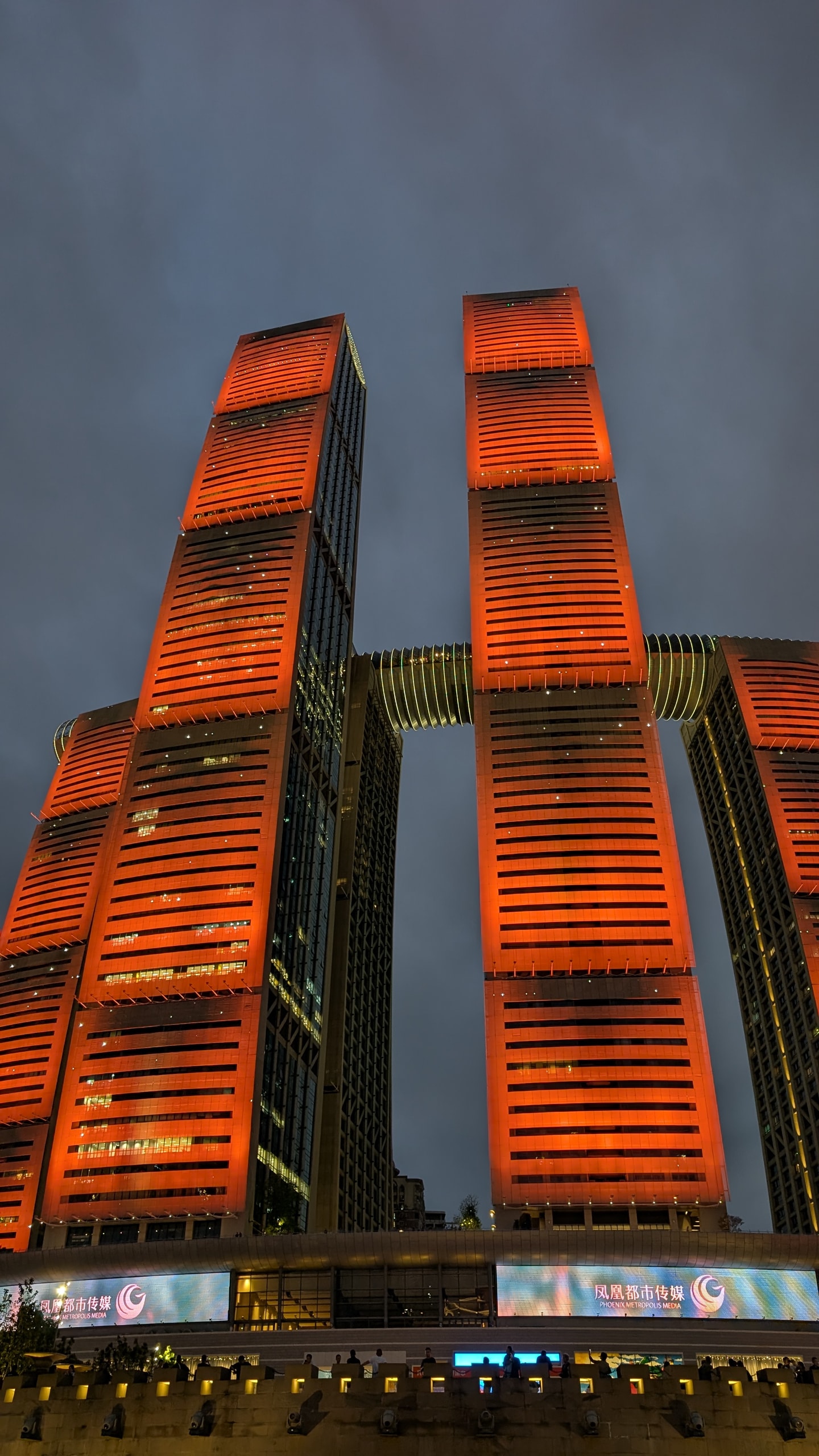 Raffles City Chongqing towers glowing orange at night with the Crystal skybridge connecting them above silhouetted viewers
