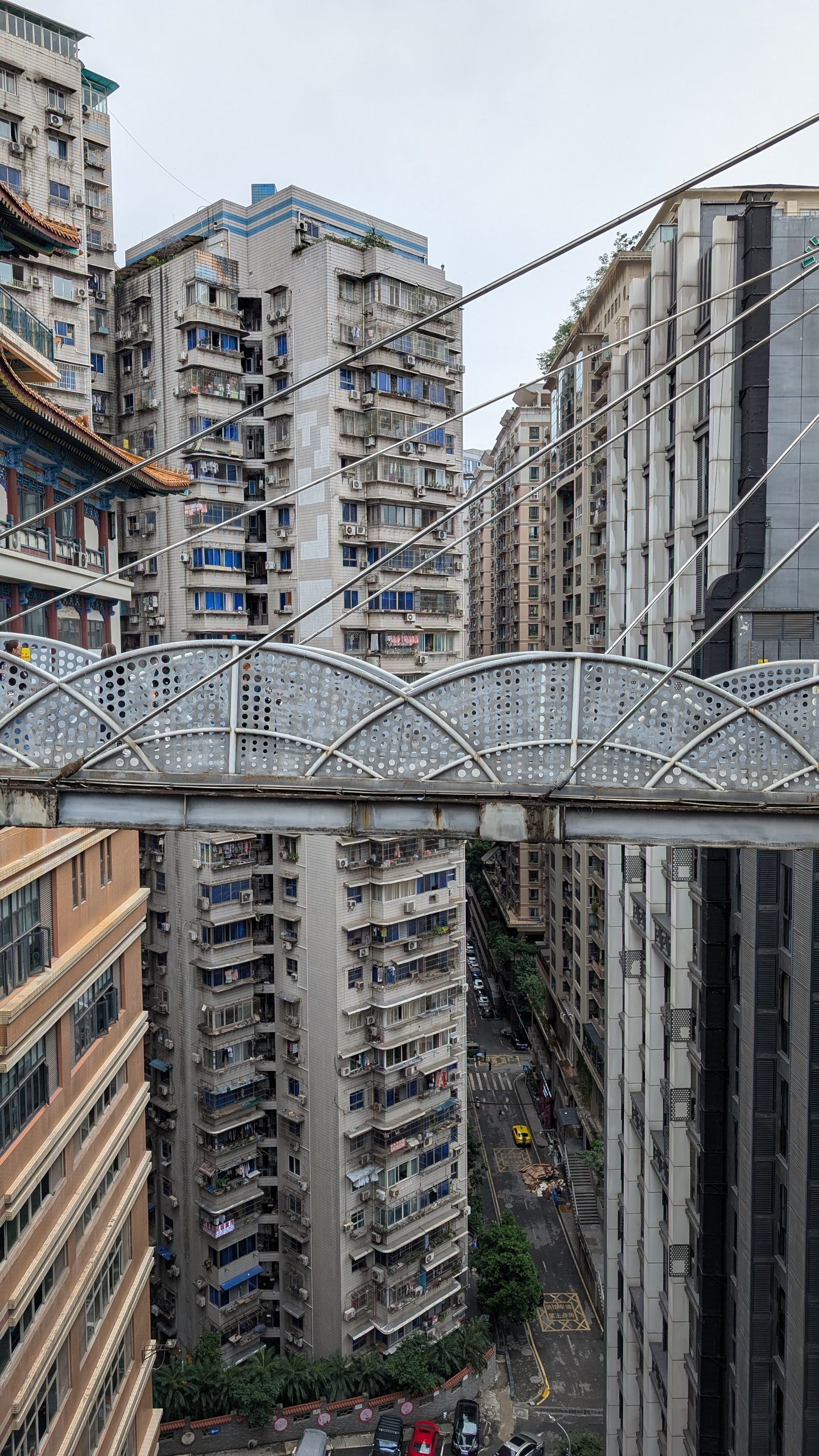 Looking down from a high pedestrian bridge between Chongqing apartment towers, with a yellow taxi on the street far below