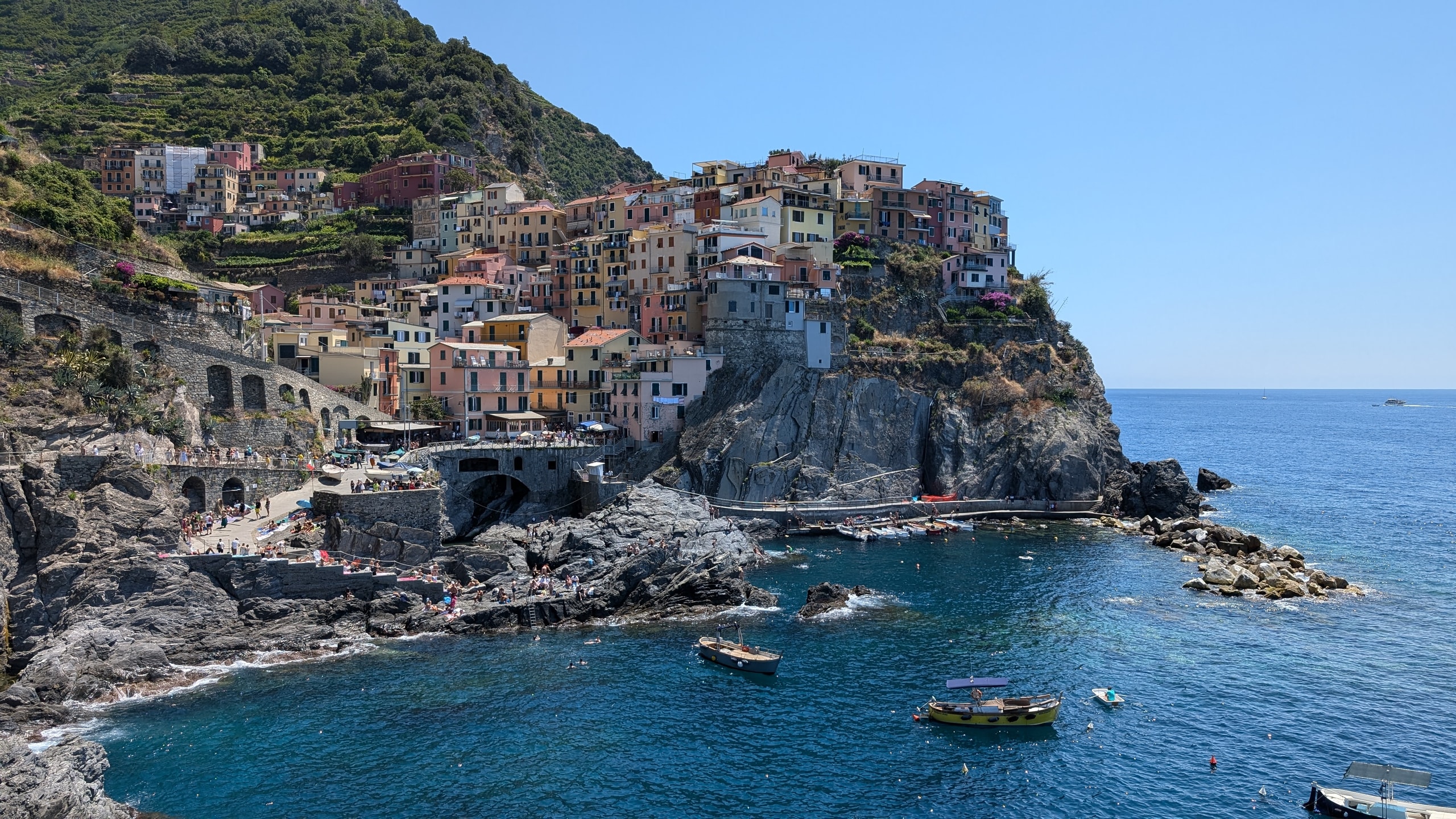 Pastel houses of Manarola stacked on a dark rock headland above a blue cove with small boats and terraced hills behind