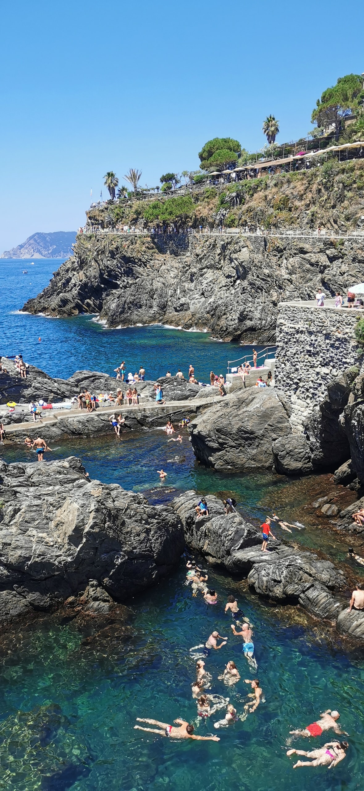Swimmers floating in clear green water between dark rocks in Manarola, with people sunbathing on a concrete landing
