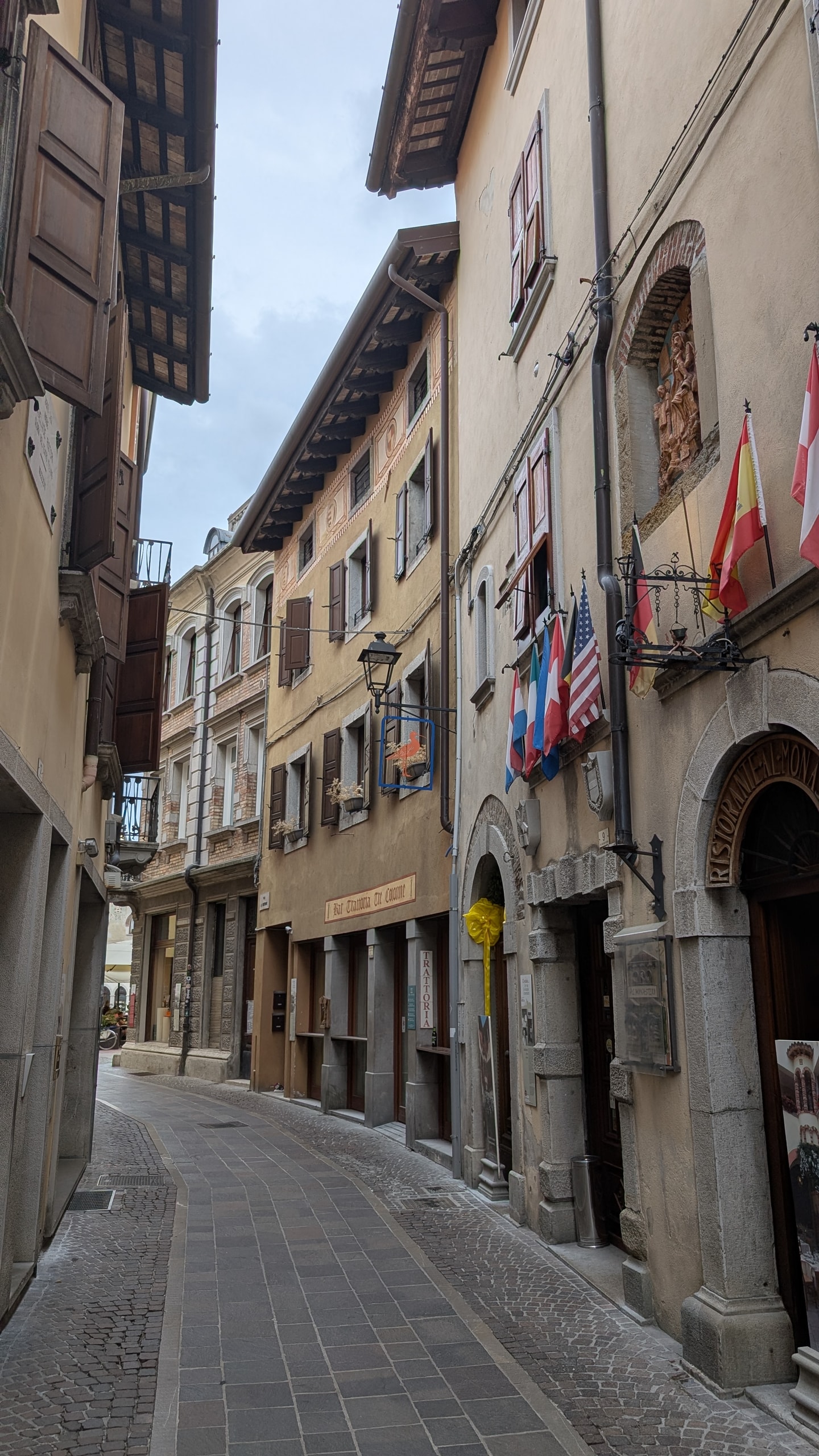 A narrow stone-paved street in Cividale del Friuli lined with old plaster houses, timber eaves, wooden shutters and flags outside a trattoria