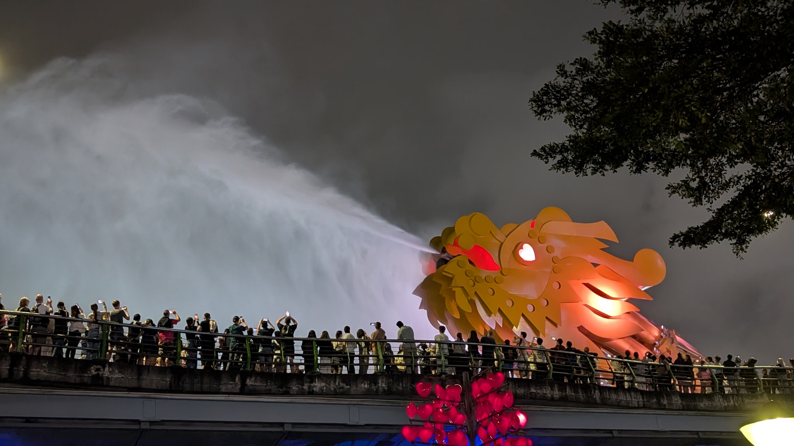 The glowing gold and red head of the Dragon Bridge spraying a huge jet of water into the night sky, with a line of people along the bridge railing filming it