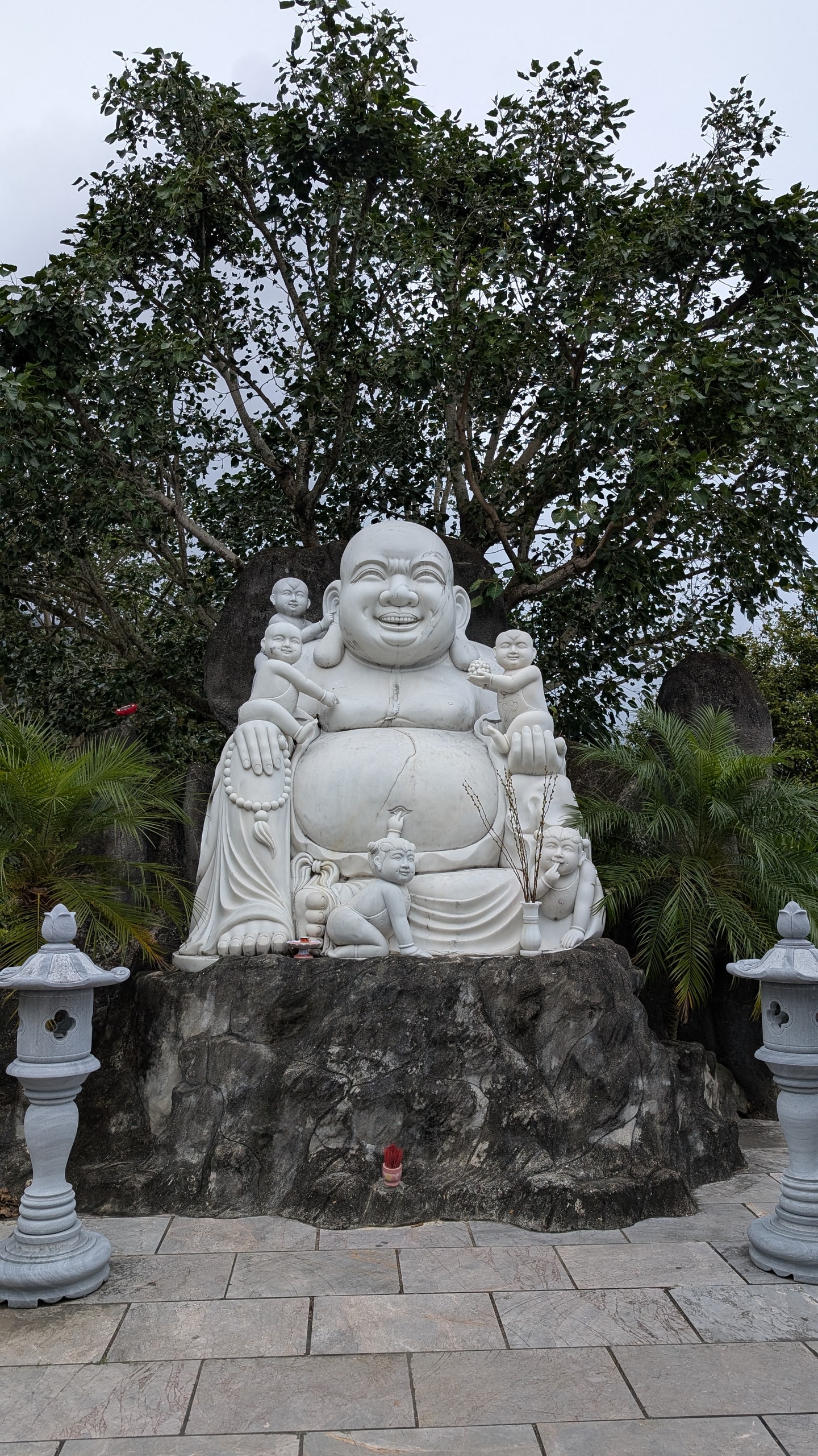 A large white marble statue of a laughing, big-bellied Buddha with five small children climbing on him, set on a dark rock with stone lanterns either side
