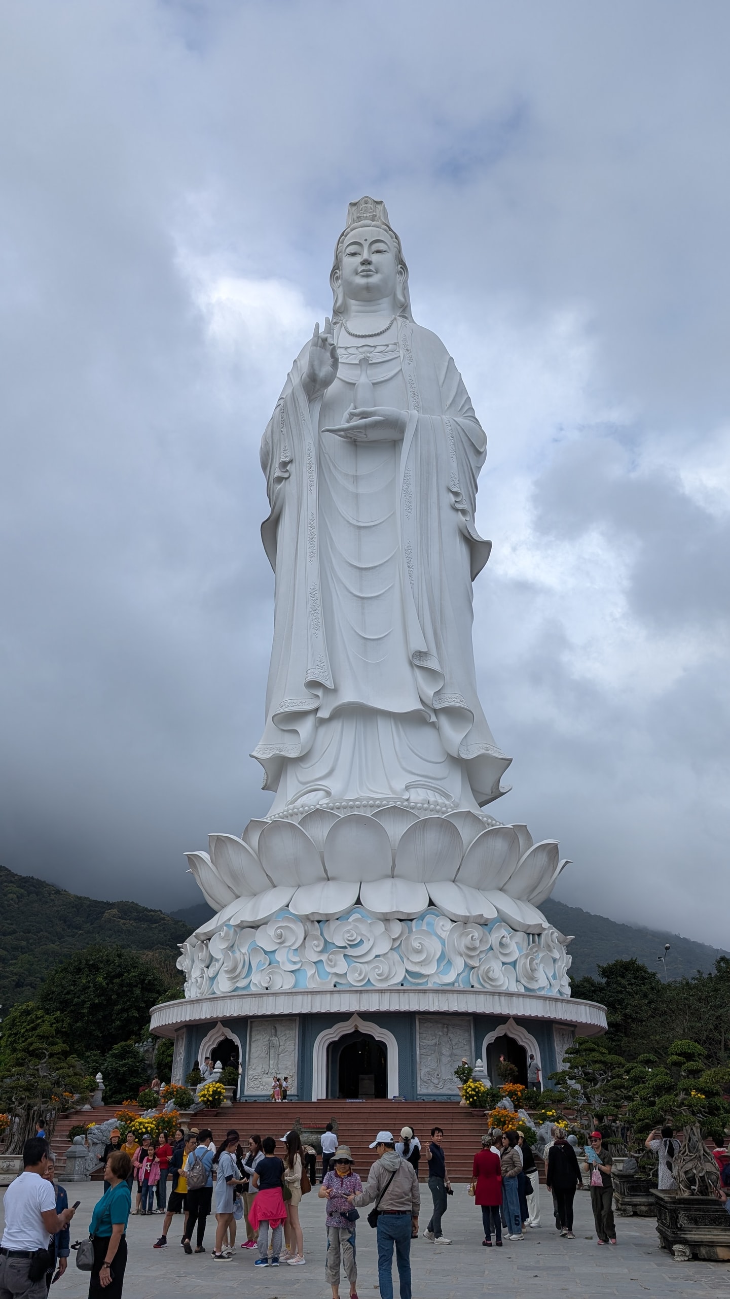 The white Lady Buddha statue at Linh Ung Pagoda standing on a giant lotus base under a cloudy sky, holding a small vase, with visitors on the plaza below