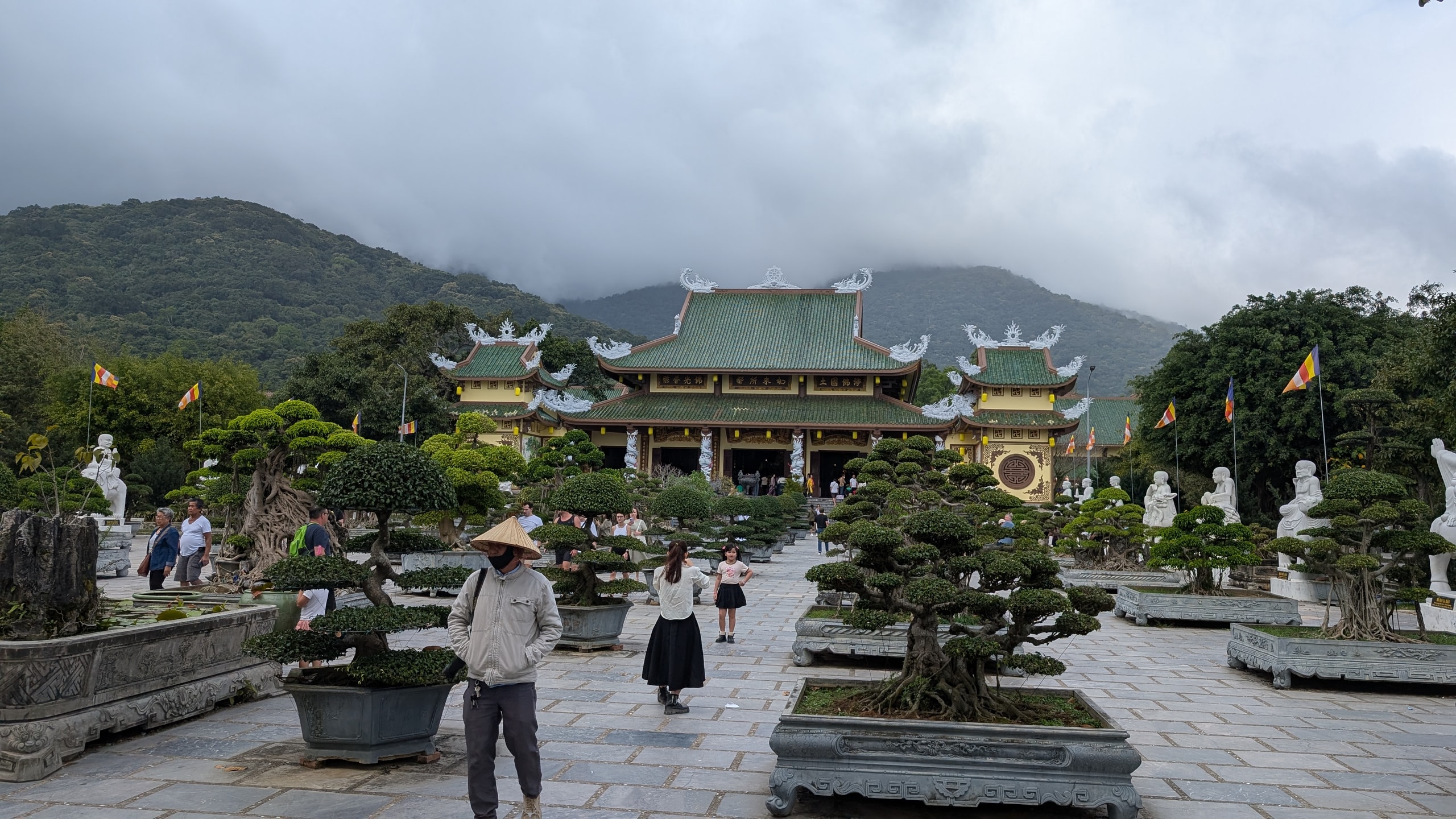 The main hall of Linh Ung Pagoda with green tiled roofs, framed by large bonsai trees in stone planters and white statues, with low cloud sitting on the forested mountain behind