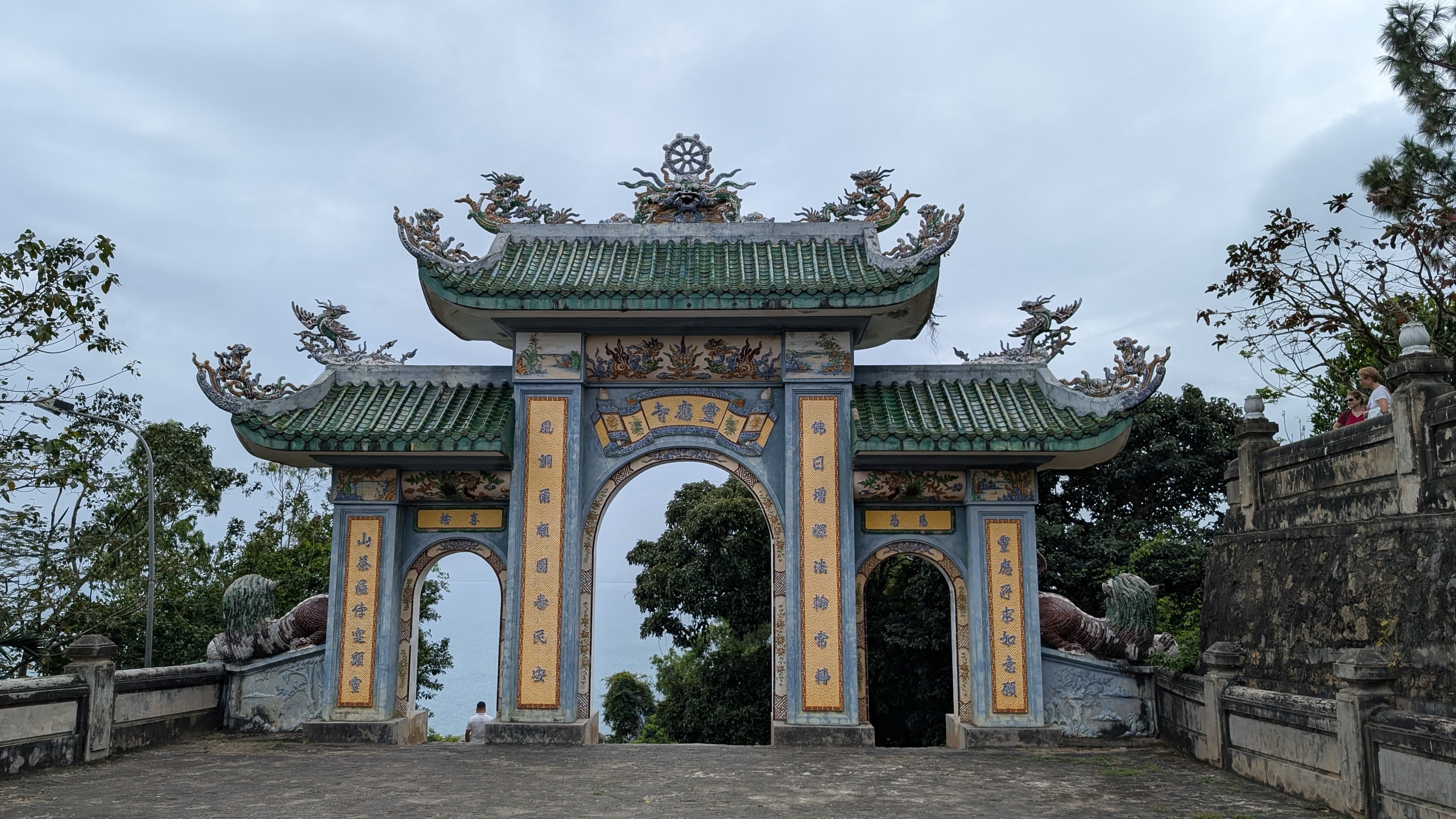 A three-arched temple gate at Linh Ung Pagoda with green tiled roofs, dragon decorations and yellow panels of Chinese characters, with the grey sea visible through the arches