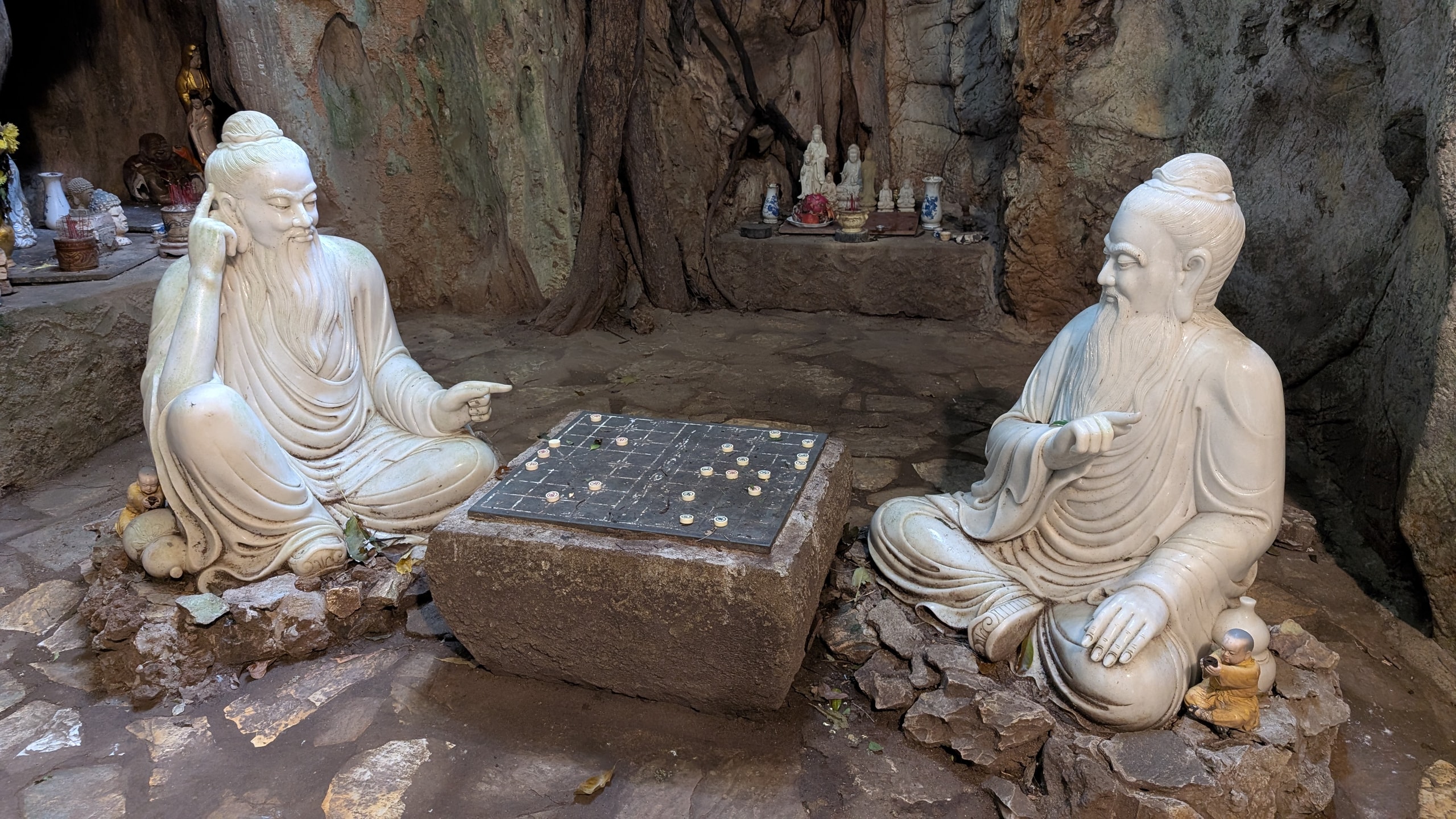 Two white marble statues of bearded old men sitting cross-legged on either side of a stone table with a Chinese chess board, in a rock grotto with small altars behind