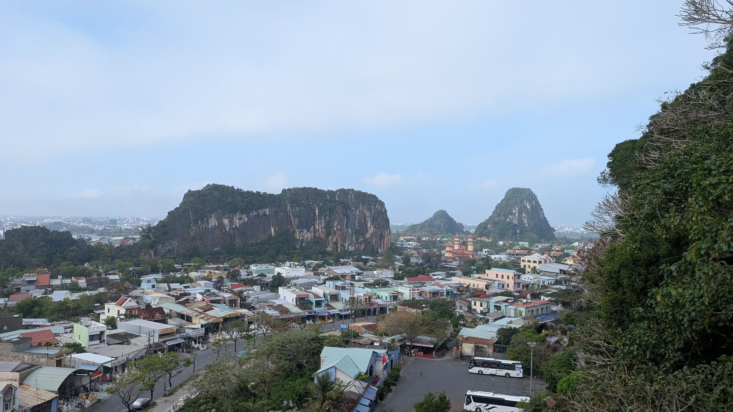 View from the top of Thuy Son over rooftops, a road lined with stone-carving shops and a car park with two coaches, towards three steep limestone hills rising out of the town