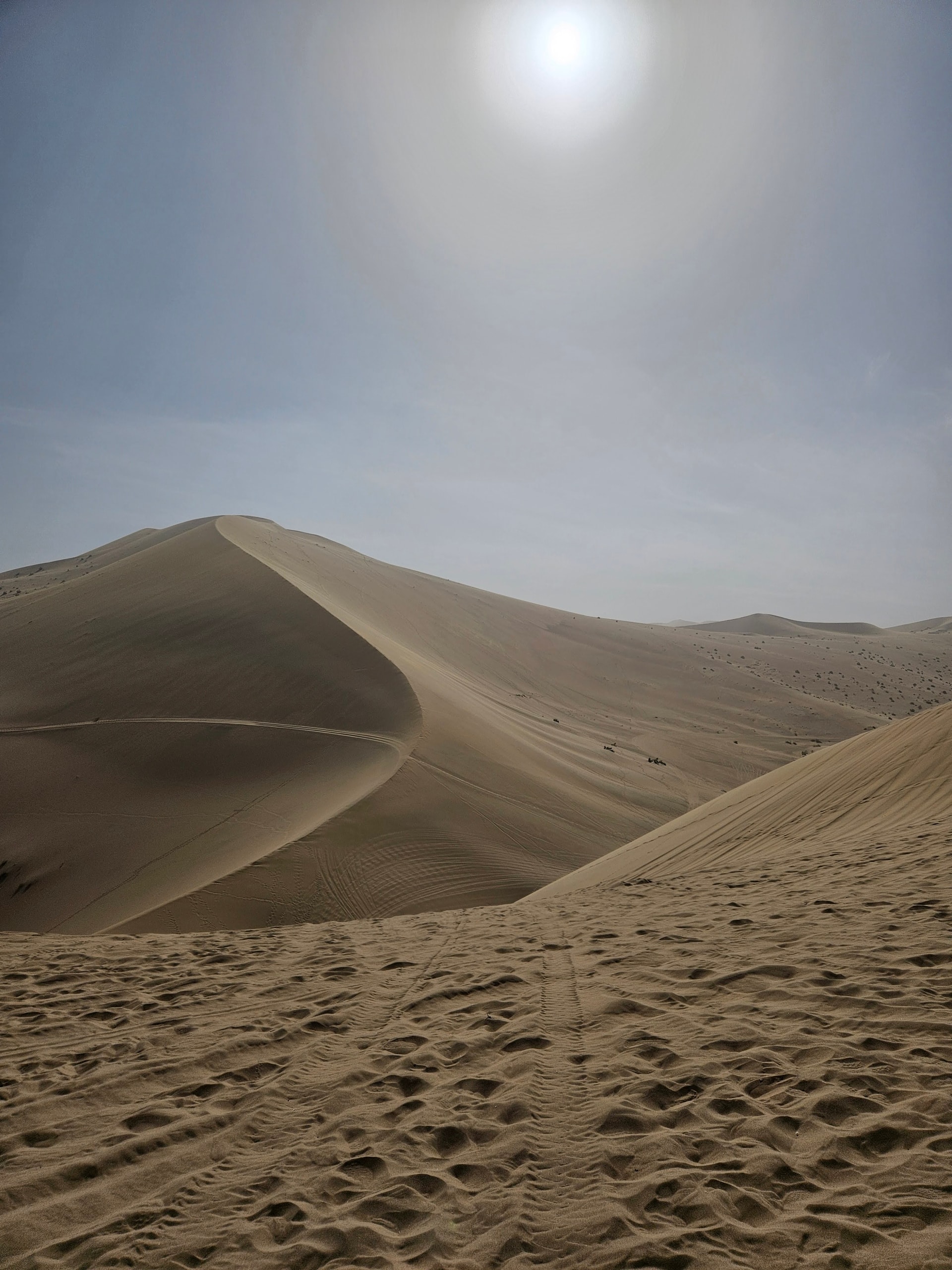 Rolling sand dunes near Dunhuang with sharp crests, sparse desert shrubs, and footprints and tire tracks in the foreground sand