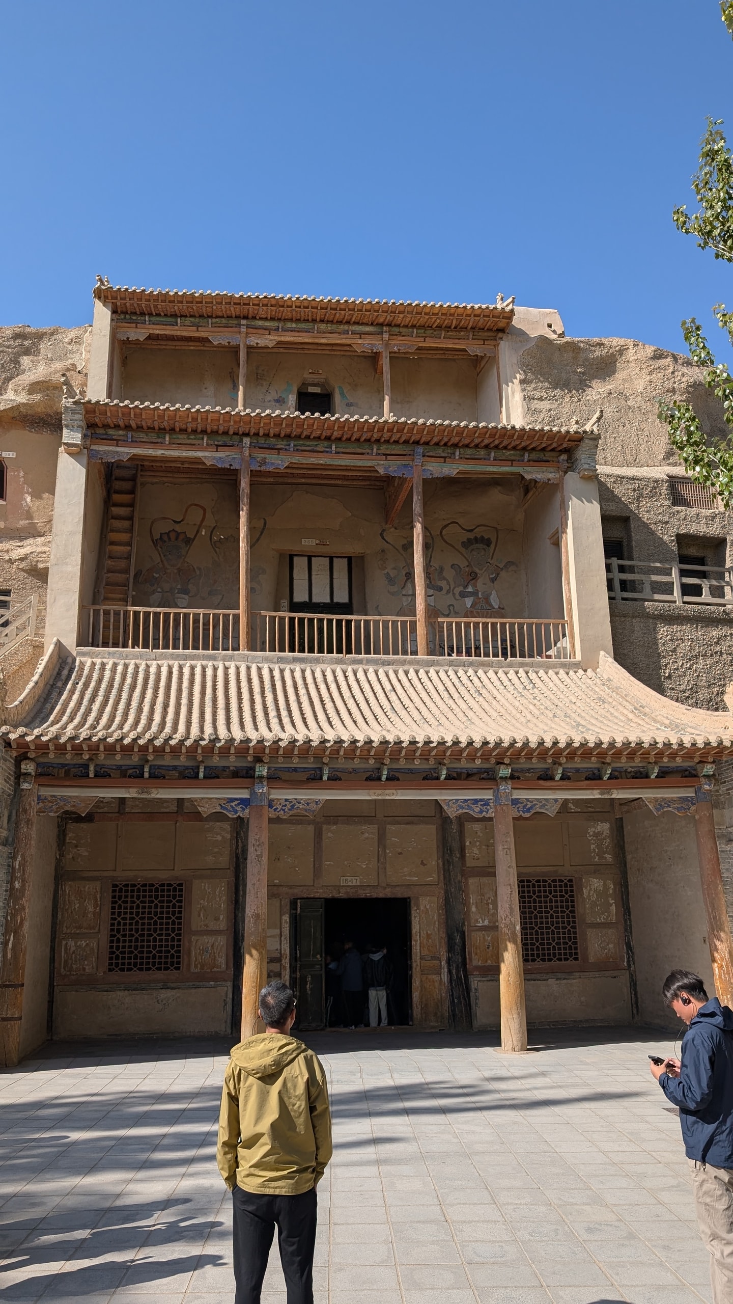 A three-tier wooden temple facade built into the Mogao cliff face with painted eaves, lattice windows, and visitors in the shaded doorway