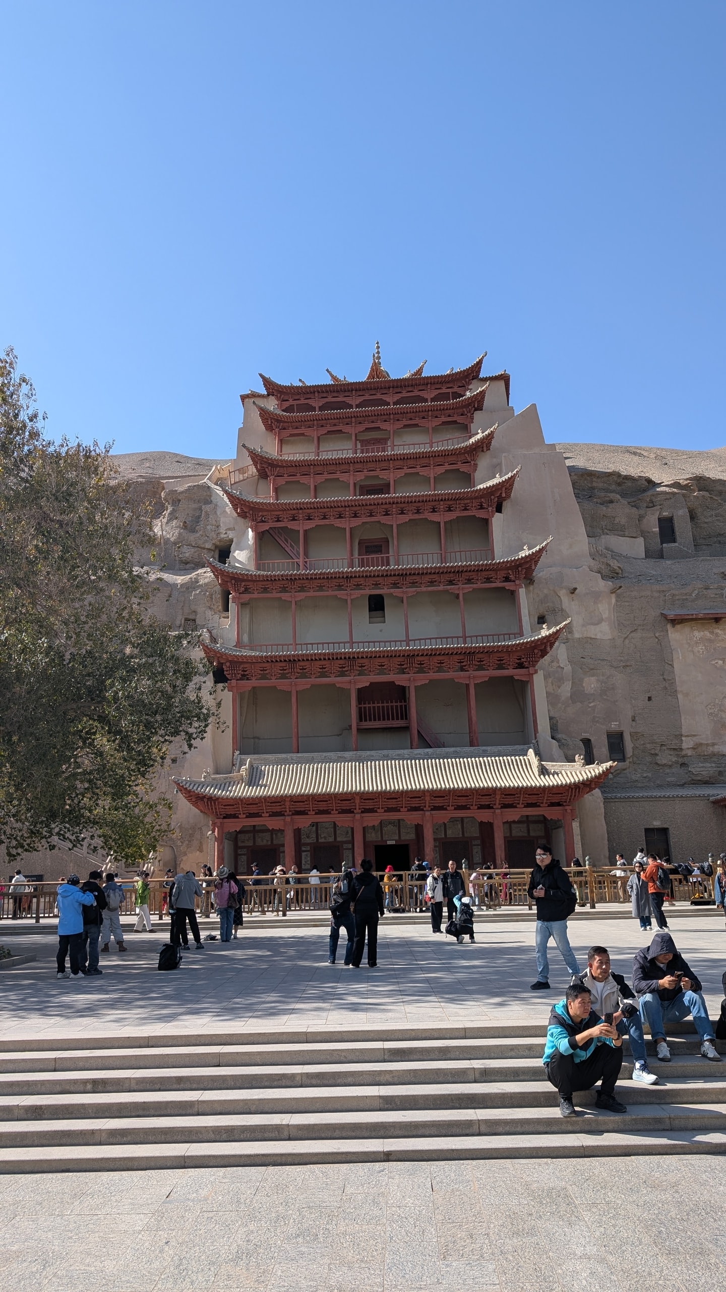 The nine-story wooden facade of Cave 96 at the Mogao Caves built into a sandstone cliff, with visitors in the paved courtyard below
