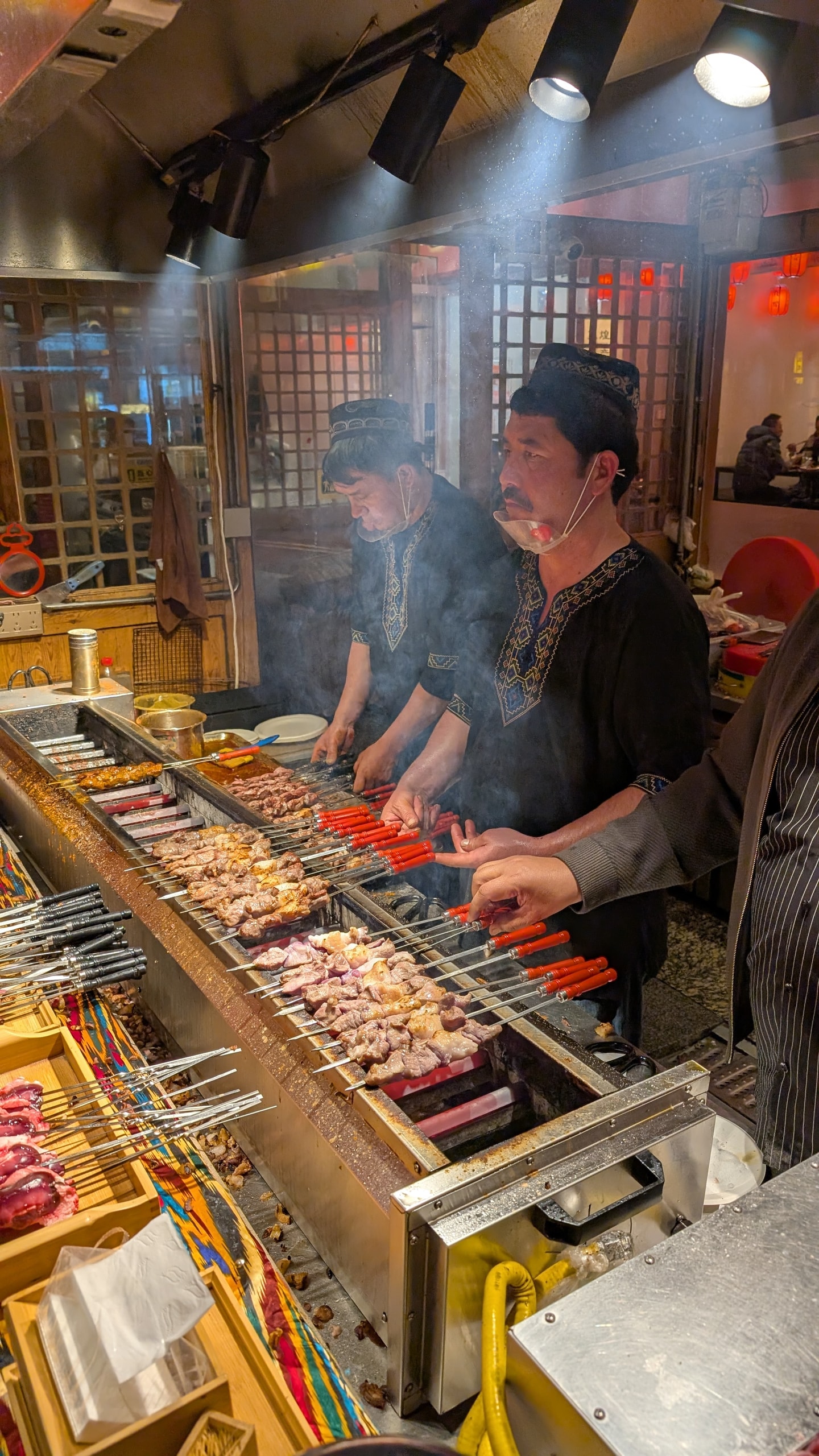 Two men in embroidered caps grilling rows of lamb skewers over charcoal at a Dunhuang night market stall, smoke rising under bright lights