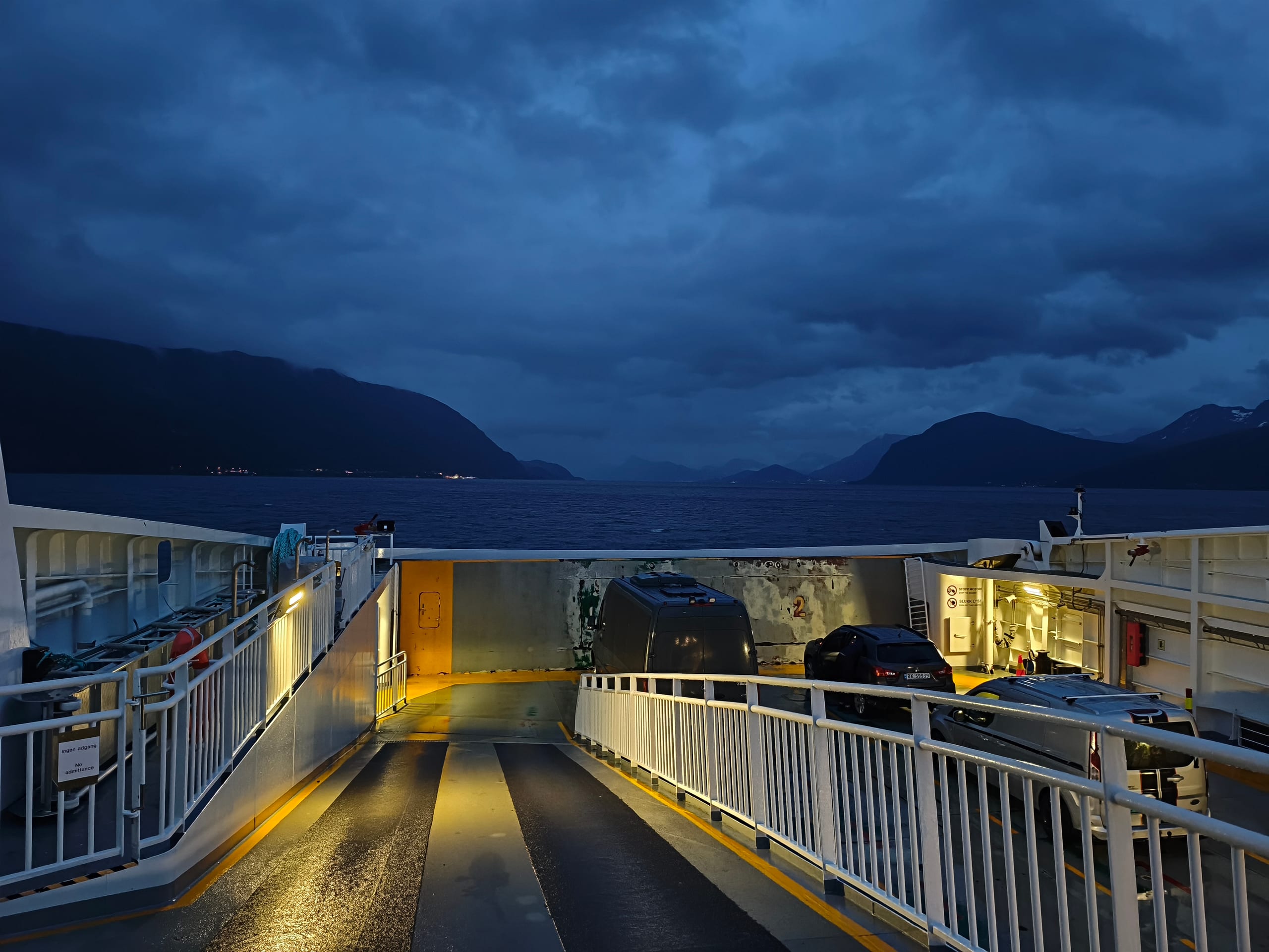 Night view from a Norwegian car ferry deck over dark fjord water with lit railings and mountain silhouettes
