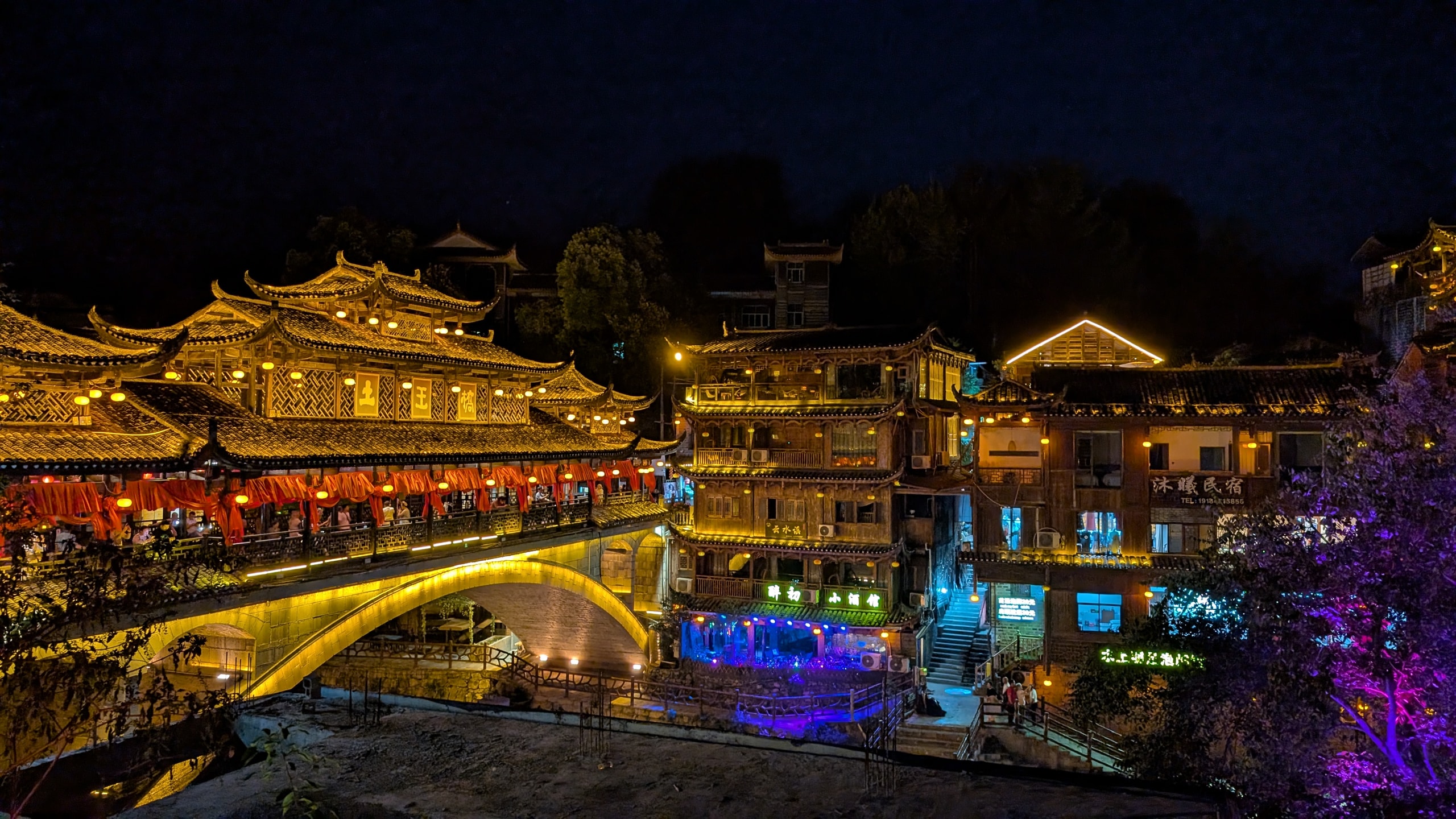 Illuminated covered bridge and multi-story wooden buildings of Furong Ancient Town glowing gold against a black night sky