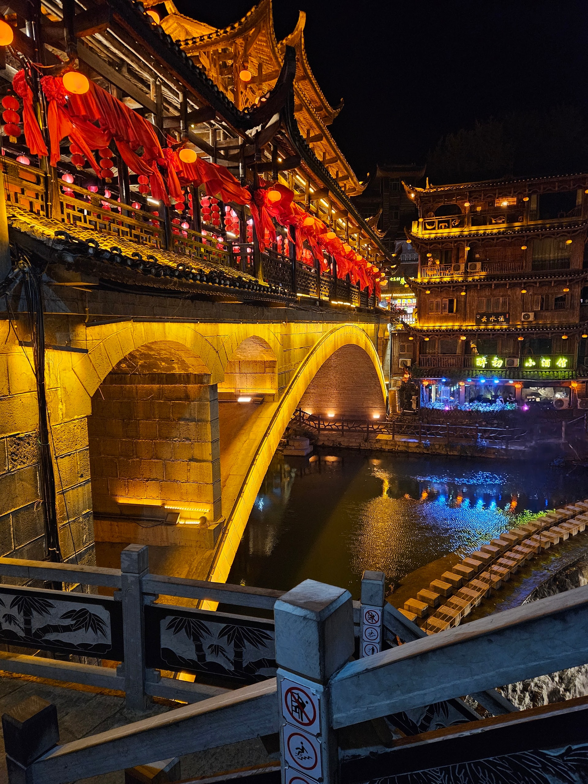Stone arch bridge and lit wooden building reflected in the river at Furong Ancient Town at night