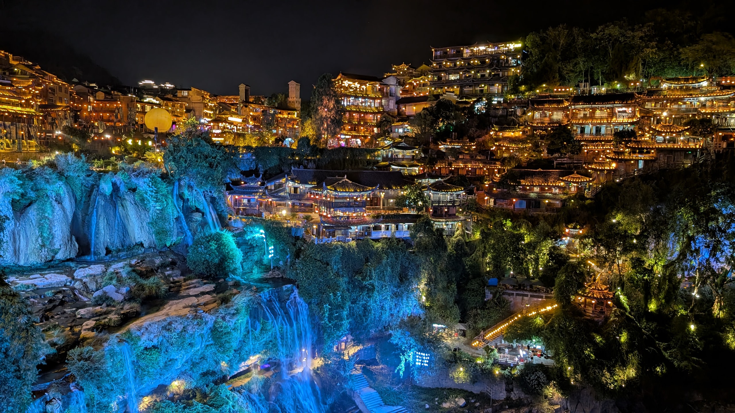 Furong Ancient Town at night with golden-lit wooden houses stacked above a blue-illuminated waterfall