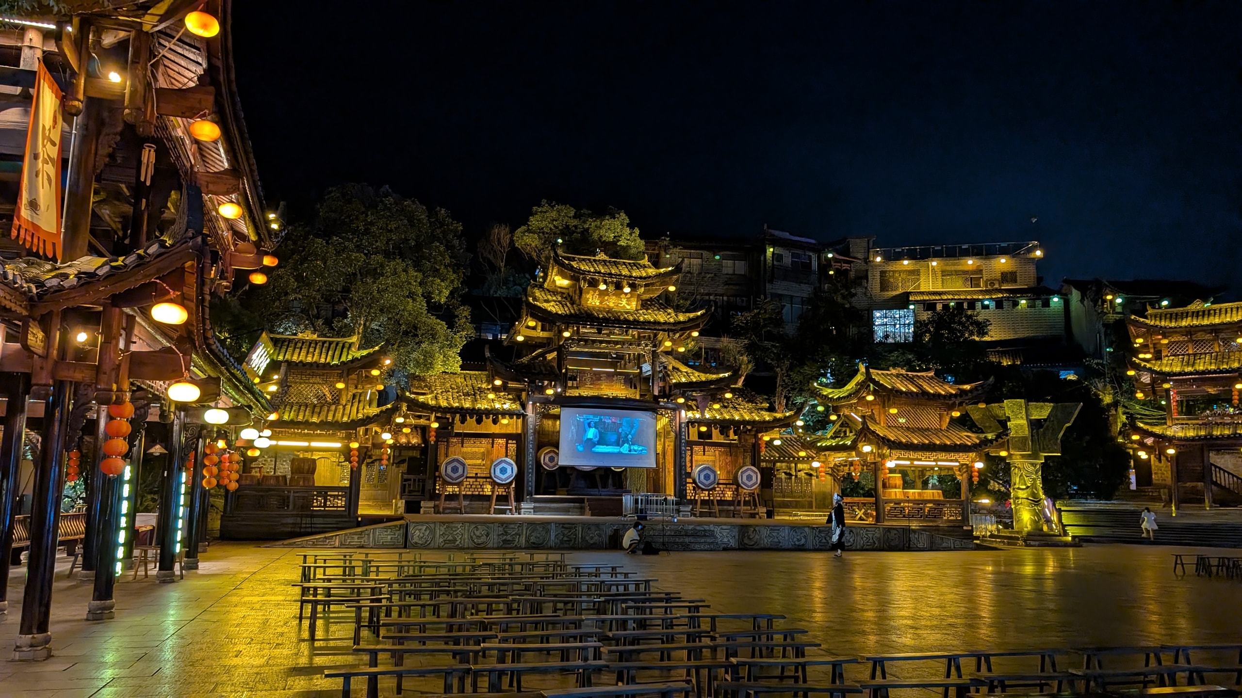 Night plaza in Furong Ancient Town with wooden benches facing an illuminated stage, drums, and traditional multi-tier roofs