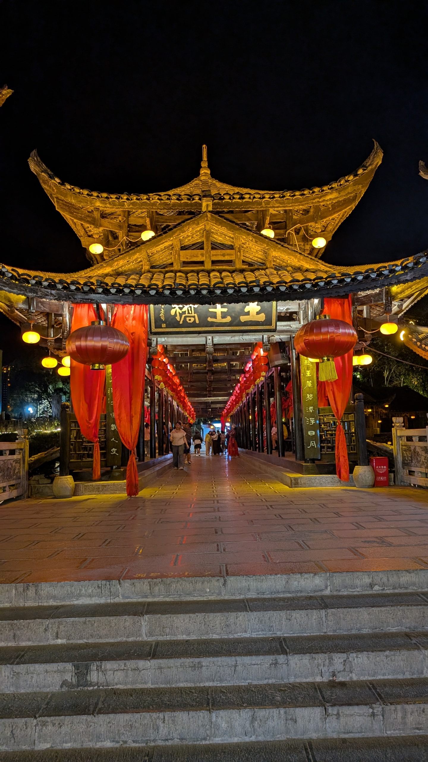 Entrance to Tuwang Bridge at night with multi-tier wooden roofs, red lanterns, and red fabric drapes over stone steps