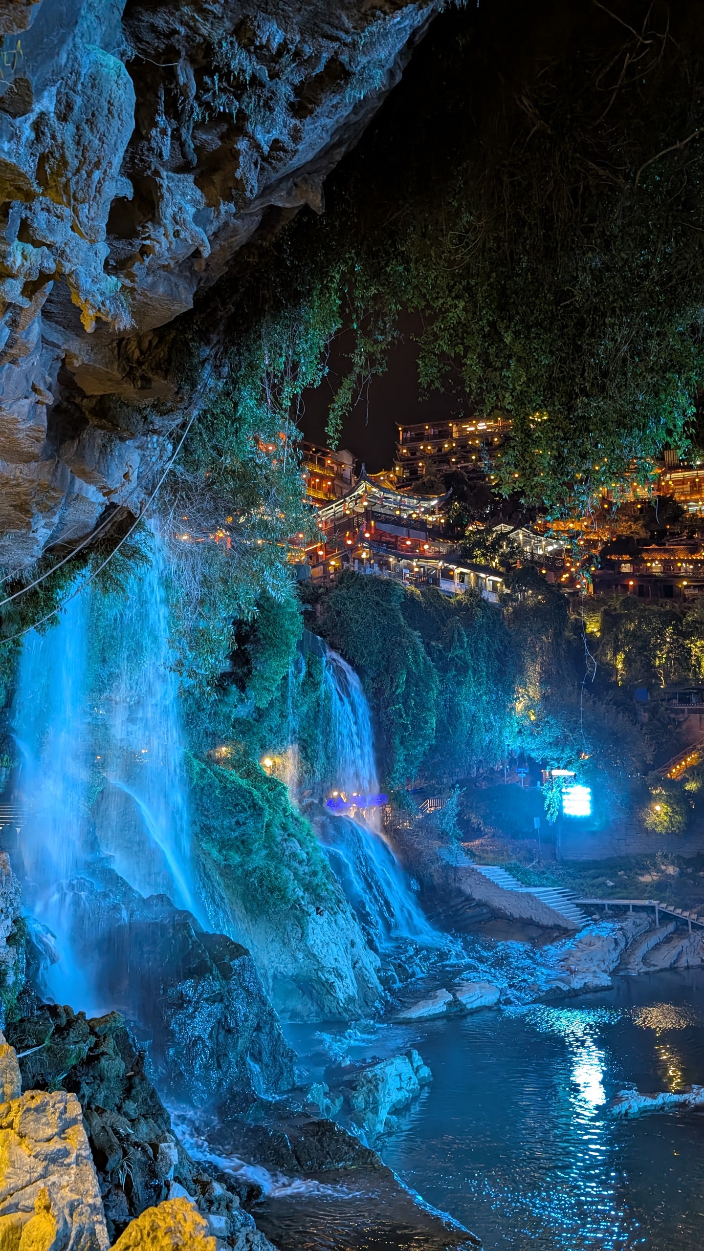 Blue-lit waterfall and golden wooden rooftops of Furong framed by a dark rock overhang and hanging vines