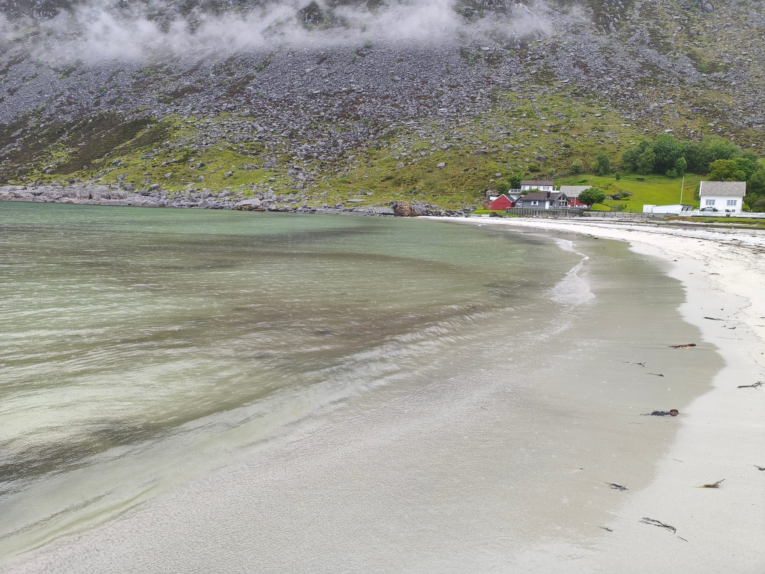 Pale sand and shallow turquoise water at Grotlesanden beach with a mist-covered mountain rising behind red boathouses