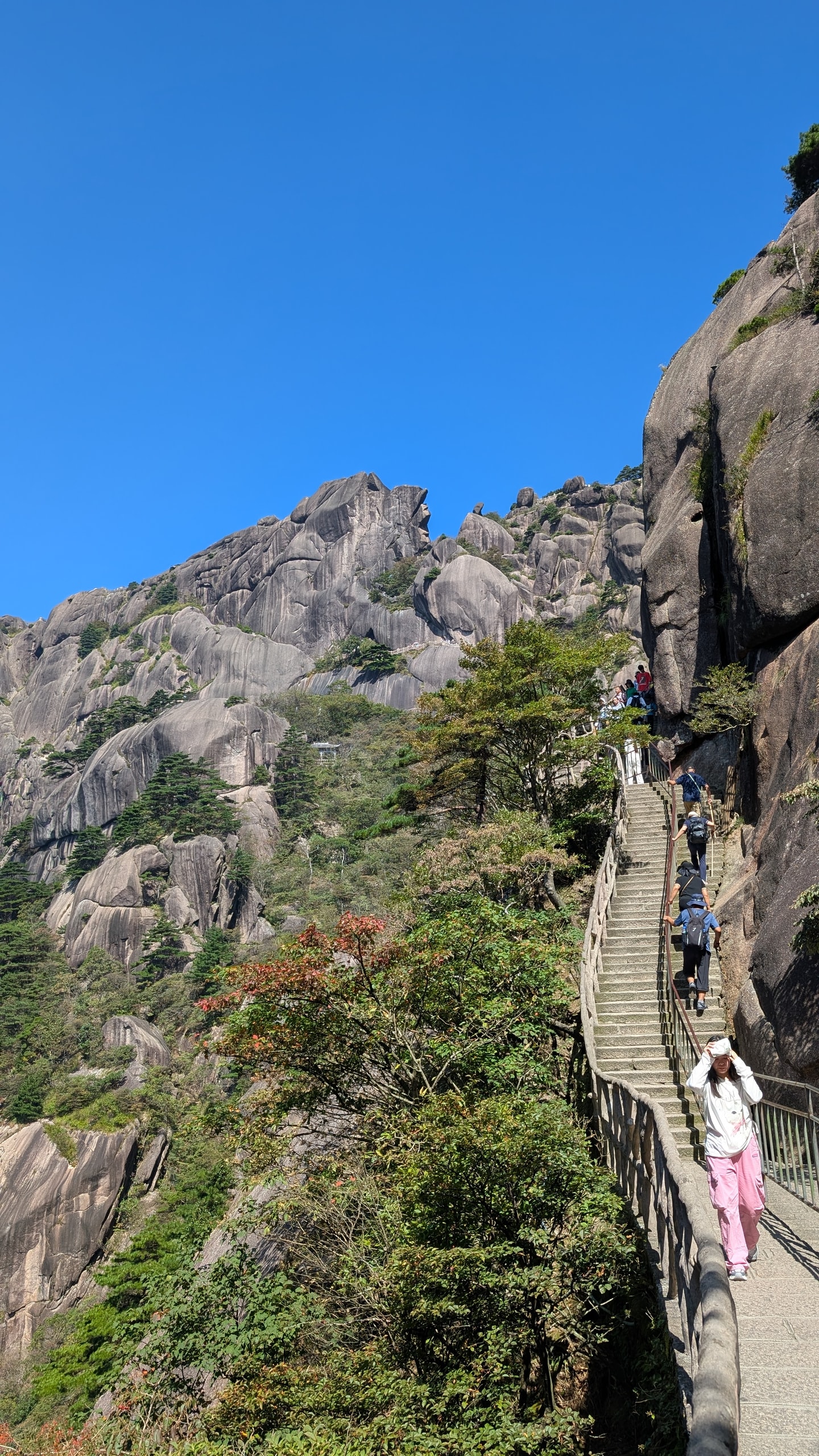 Hikers on a narrow stone staircase built into a Huangshan cliff face under a clear blue sky