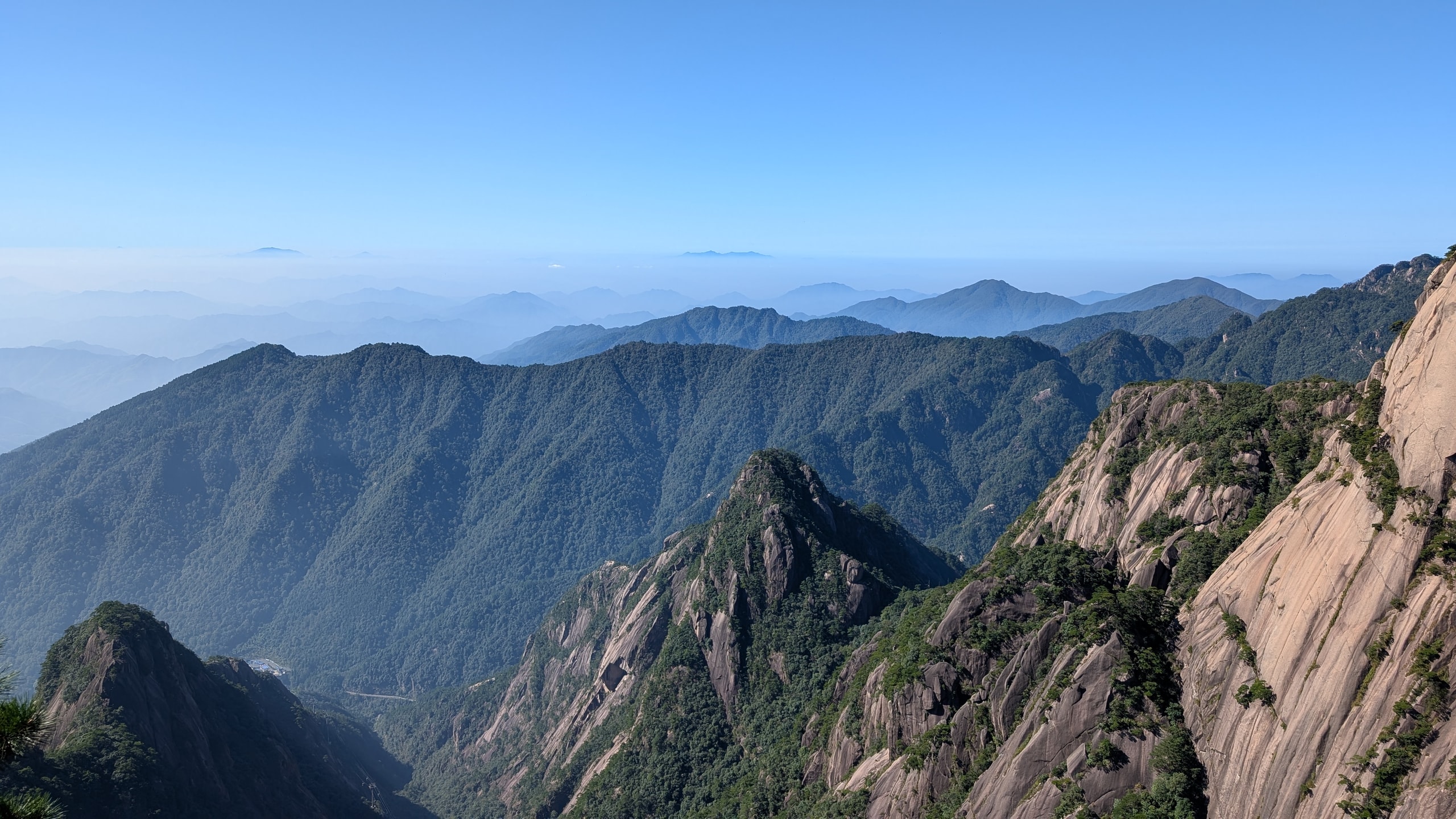 Wide view across Huangshan granite peaks and forested valleys with a cable line visible against a sheer rock face