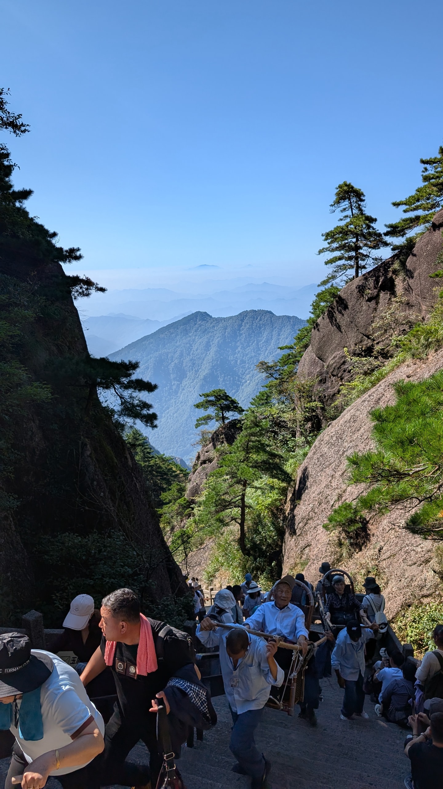 Hikers and sedan-chair porters climbing steep stone stairs between Huangshan granite cliffs with pine trees and distant blue peaks
